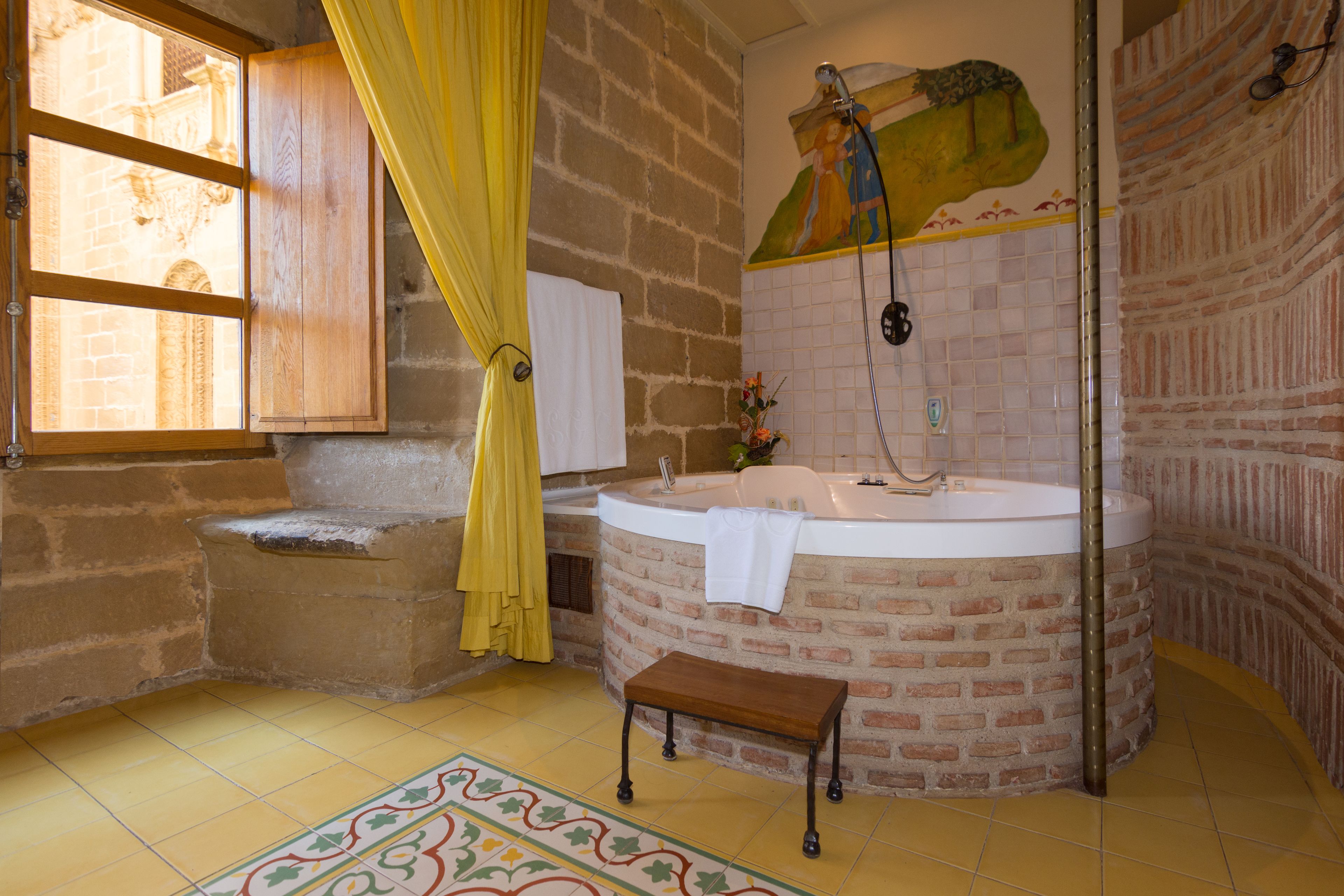 Une salle de bain avec un mur en briques, une baignoire, un lavabo, un banc et une fenêtre avec des rideaux jaunes.