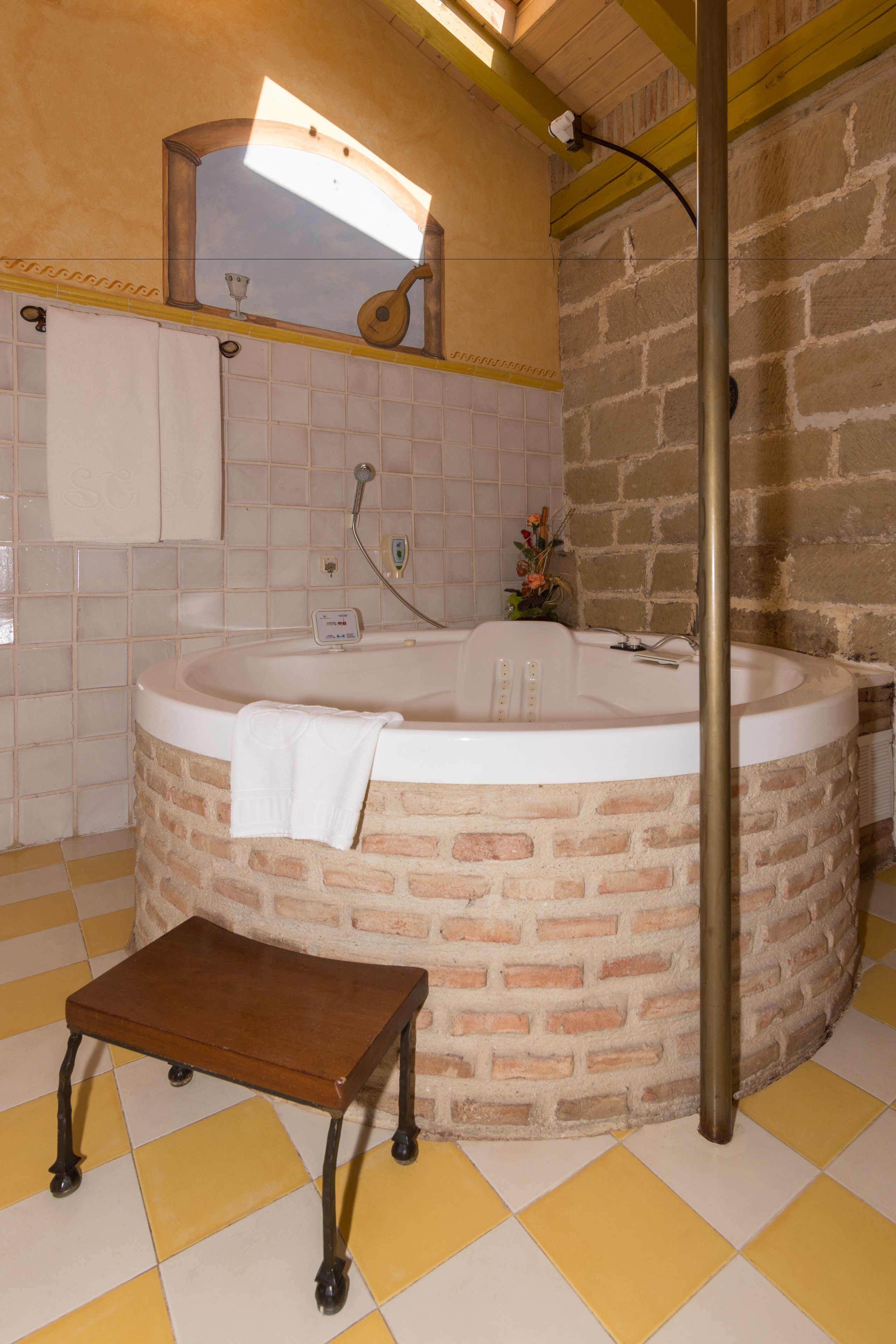 Um banheiro com parede de pedra, uma banheira, uma pequena mesa de madeira e piso quadriculado amarelo e branco.