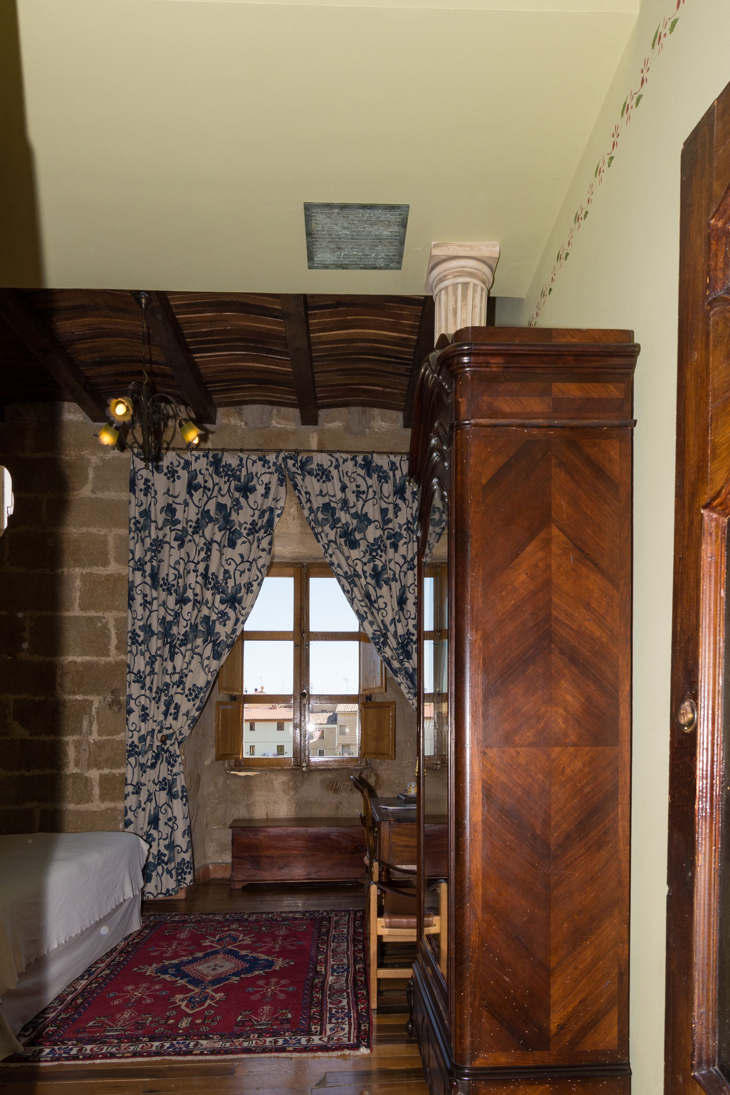 A room with a bed, a wooden wardrobe, a window with curtains, a rug, and a stone wall.