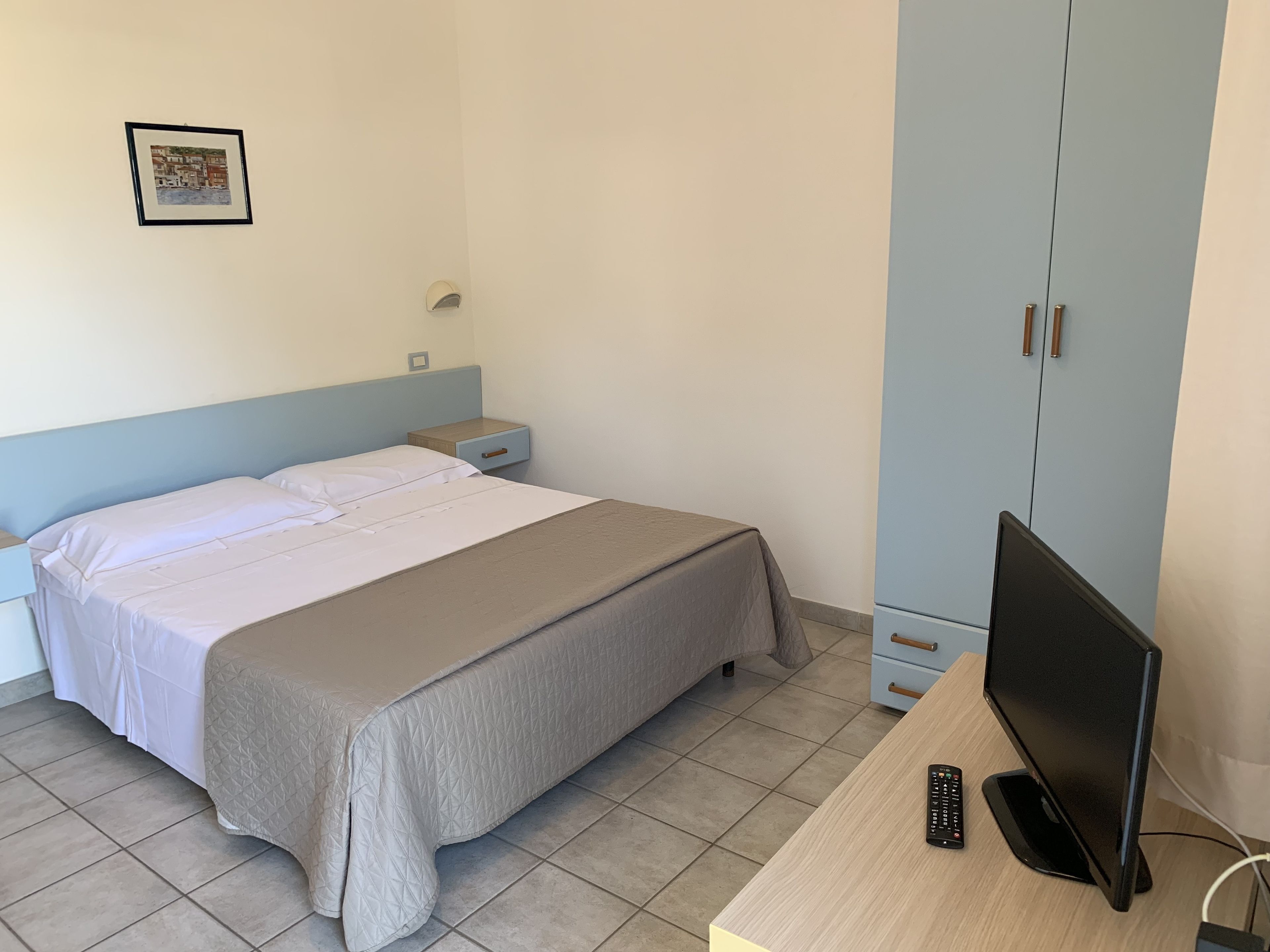 A bedroom with a bed, nightstand, wardrobe, and a desk with a television and remote control.
