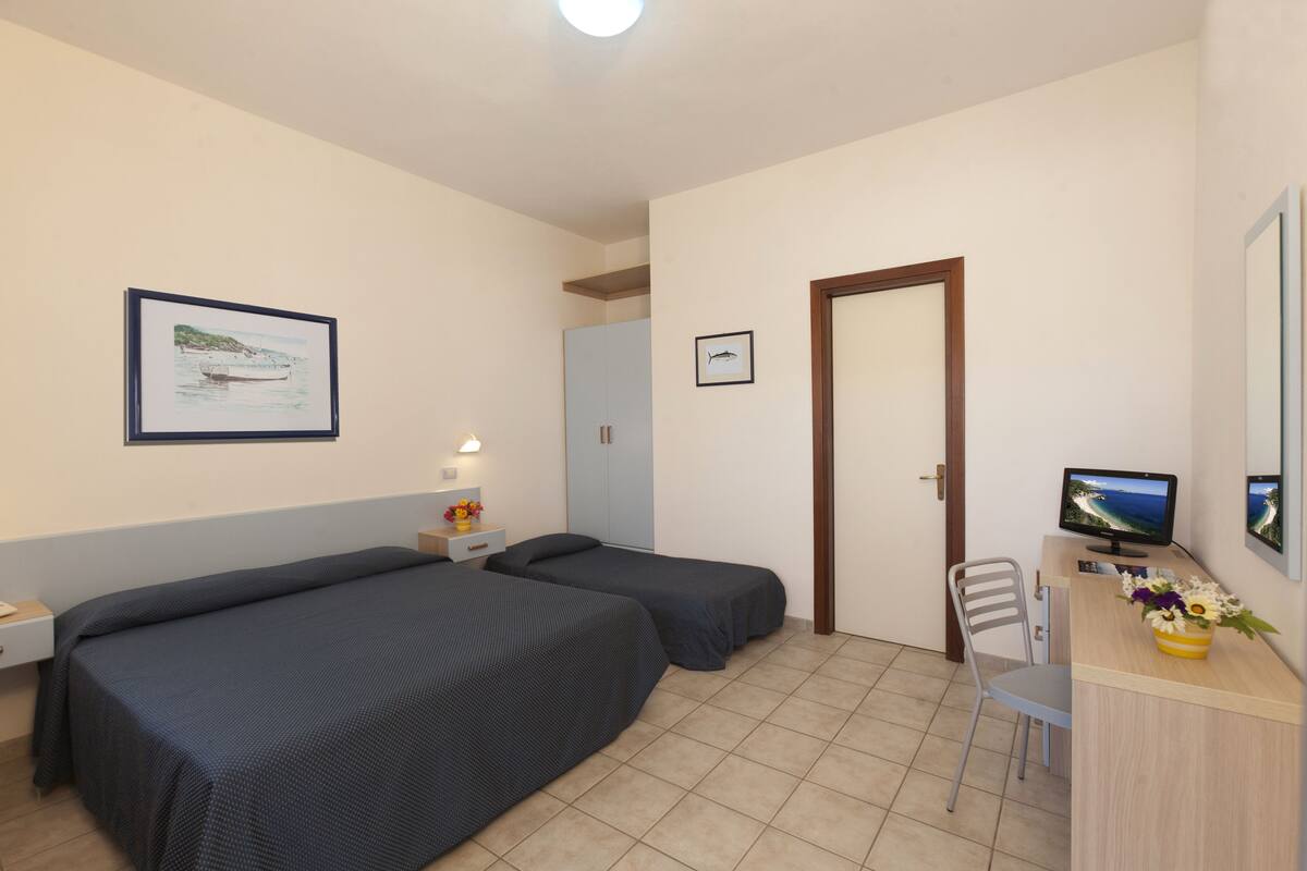 Una habitación con dos camas individuales, un escritorio con computadora, una silla y una puerta.