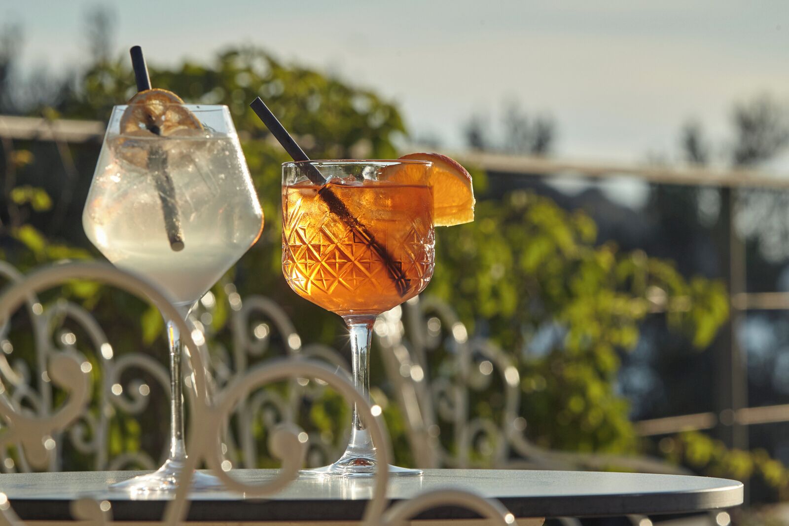 Two cocktails on a table with a metal railing and greenery in the background.
