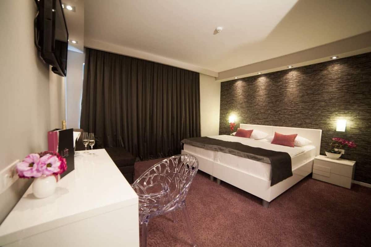 A modern hotel room with a bed, bedside table, TV, and a large window with curtains.