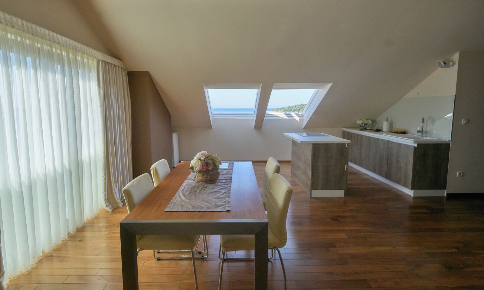A dining area with a wooden table and chairs, a flower centerpiece, and a view of the sea through skylights.