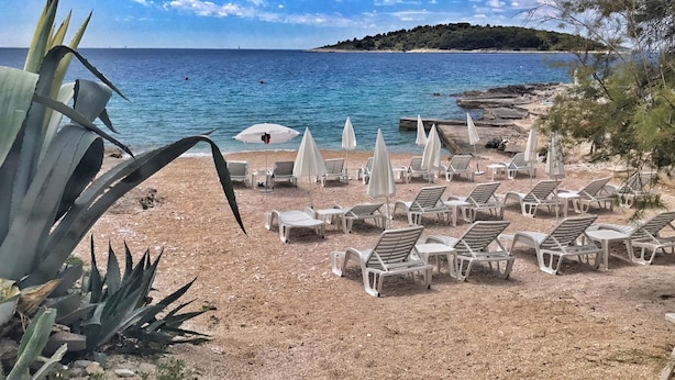 A beach with white lounge chairs and umbrellas, a rocky shoreline, and a distant tree-covered hill. - Boutique Hotel Life (Rogoznica)