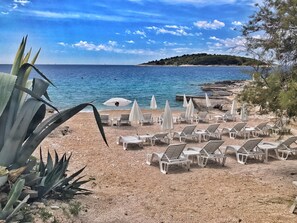 Una playa con sillas de descanso blancas y sombrillas, una costa rocosa y una colina distante cubierta de árboles.