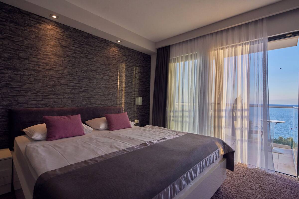 A modern bedroom with a stone accent wall, a large bed with white and purple bedding, and a view of the sea through sheer curtains.