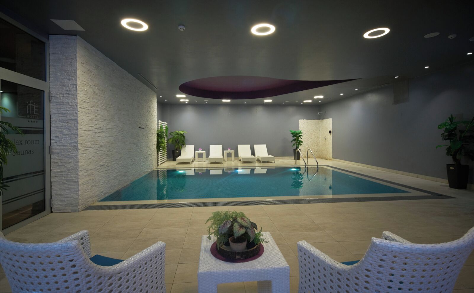 A modern indoor swimming pool with lounge chairs and a table, surrounded by stone walls and recessed lighting.