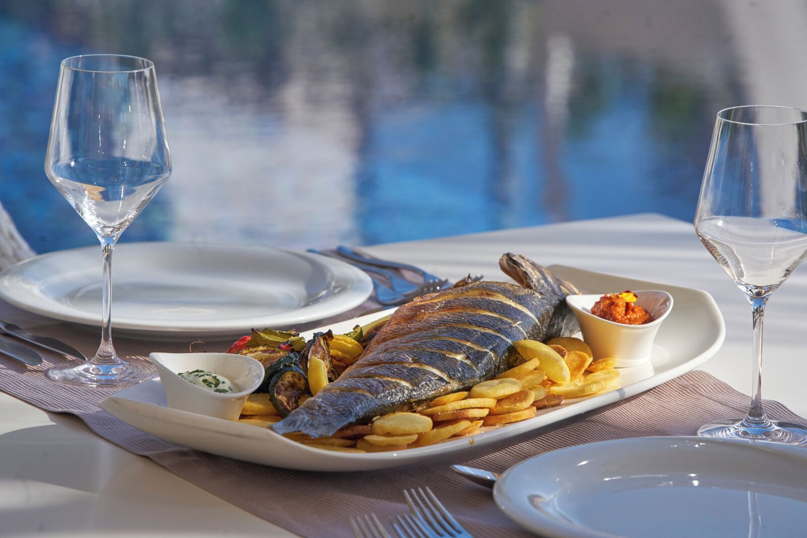 Un tavolo da pranzo con un piatto di pesce alla griglia, patatine fritte e un contorno, accompagnati da due bicchieri d'acqua.