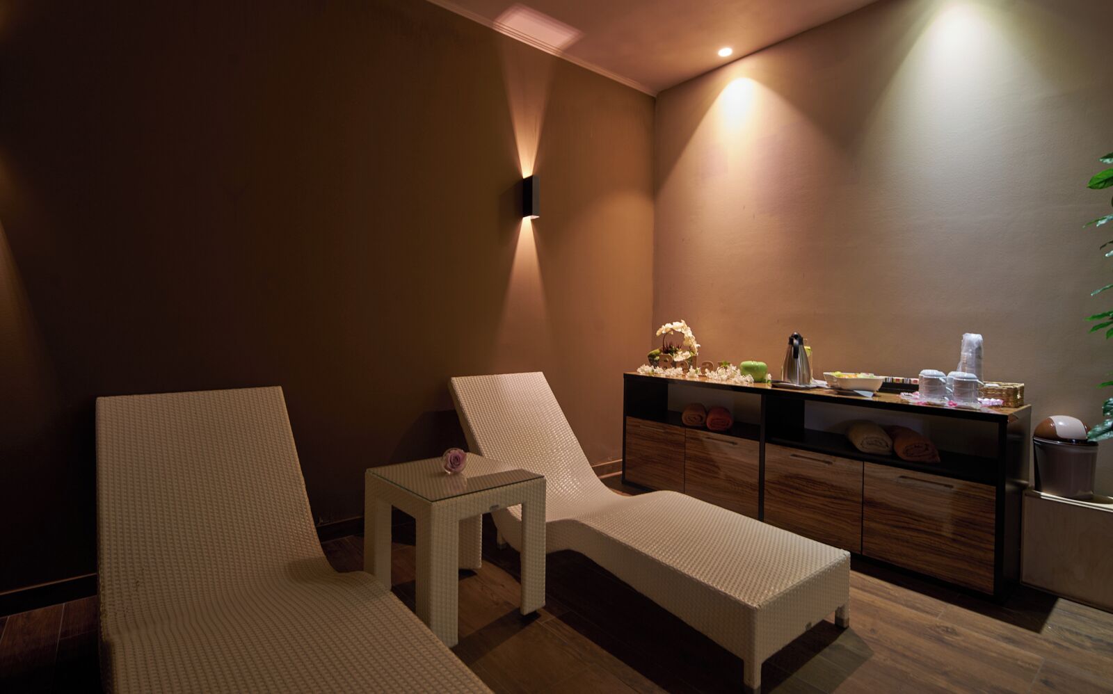 Un cuarto de spa con dos sillones blancos, una mesita con una rosa, un armario de madera con toallas y una planta.
