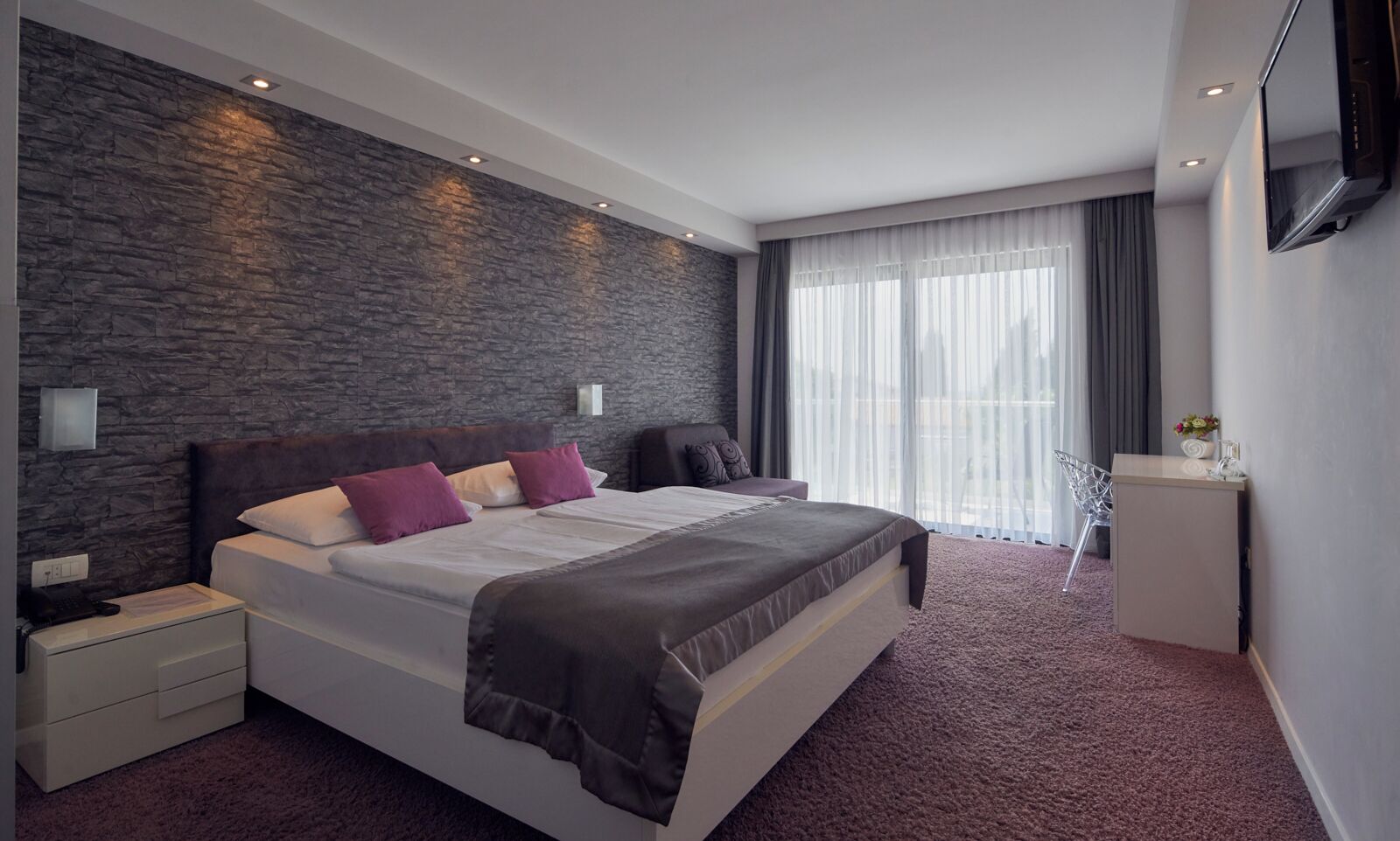 Een moderne hotelkamer met een groot bed, een accentmuur van steen, een bureau met stoel, en een groot raam met gordijnen.