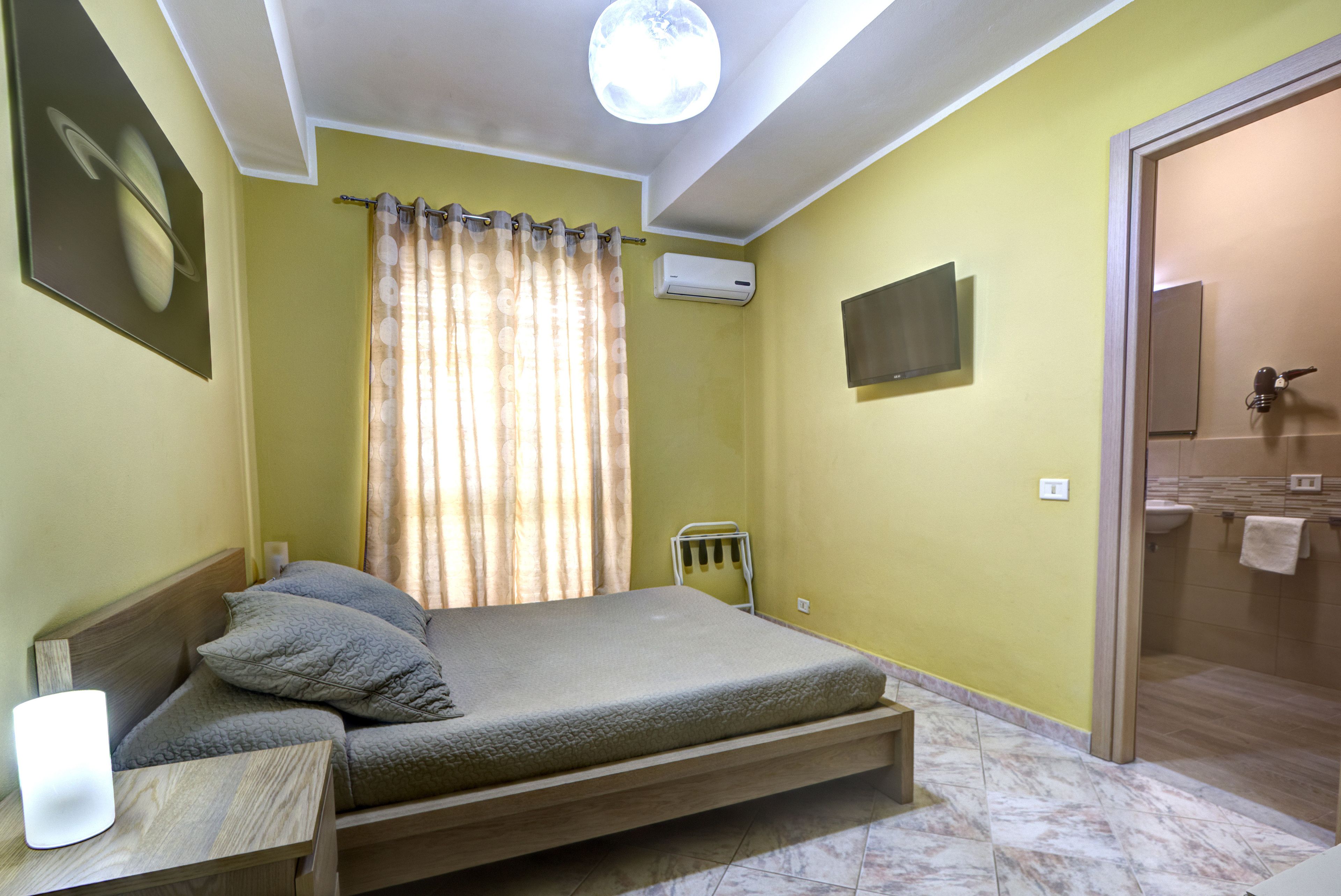 Un dormitorio con cama, una mesita de noche, ventana con cortinas, equipo de aire acondicionado, televisor montado en la pared y baño con lavamanos y toallero.