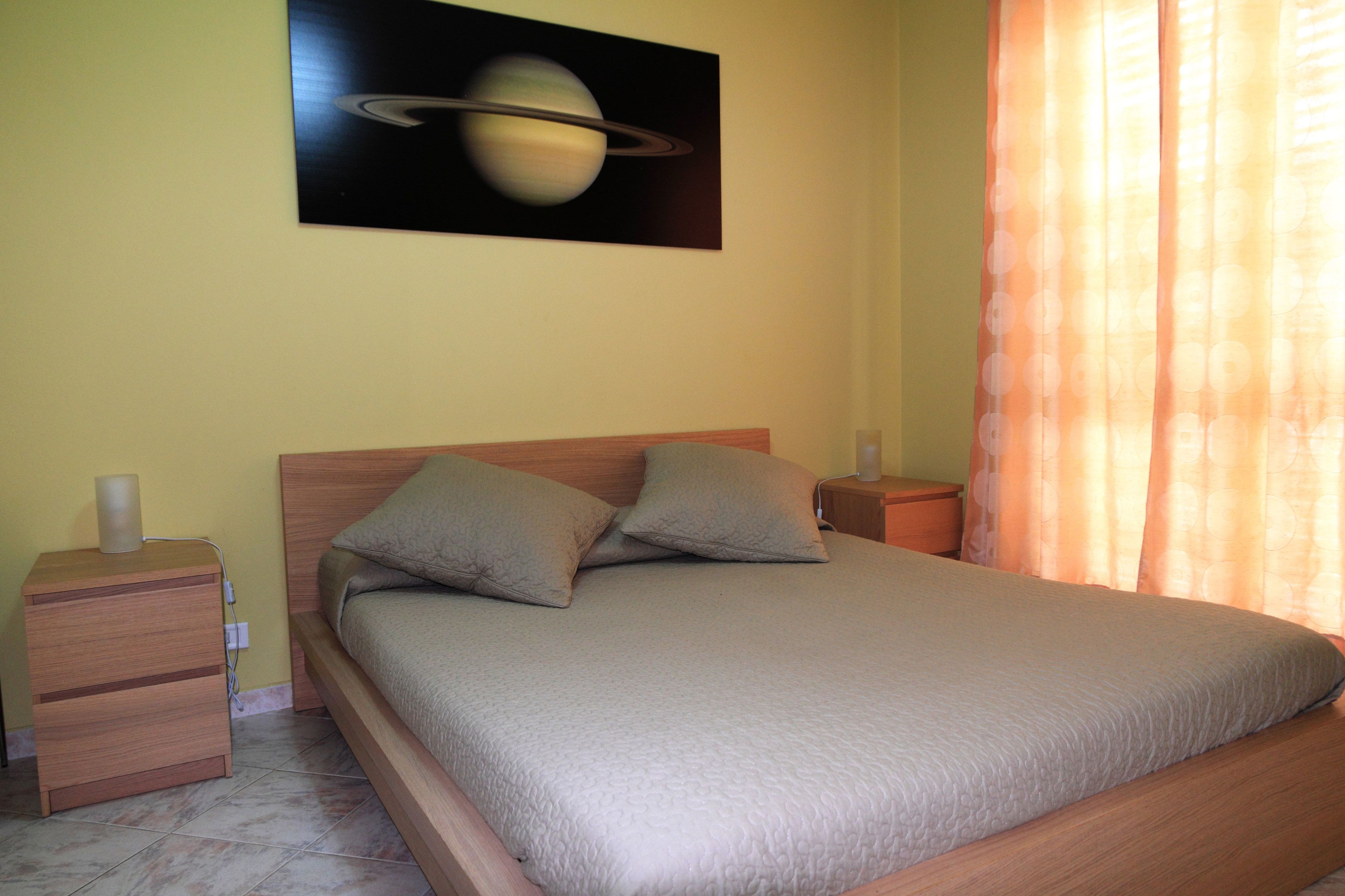 Une chambre à coucher avec un lit, deux oreillers, une table de chevet, une lampe et un tableau représentant Saturne accroché au mur.