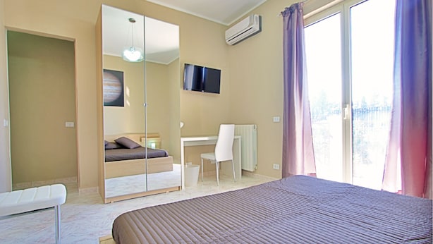 Quarto com cama, escrivaninha, cadeira, guarda-roupa e uma janela grande com cortinas.