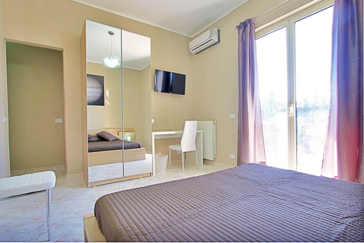 Une chambre à coucher comprenant un lit, un bureau, une chaise, une armoire et une grande fenêtre avec des rideaux.