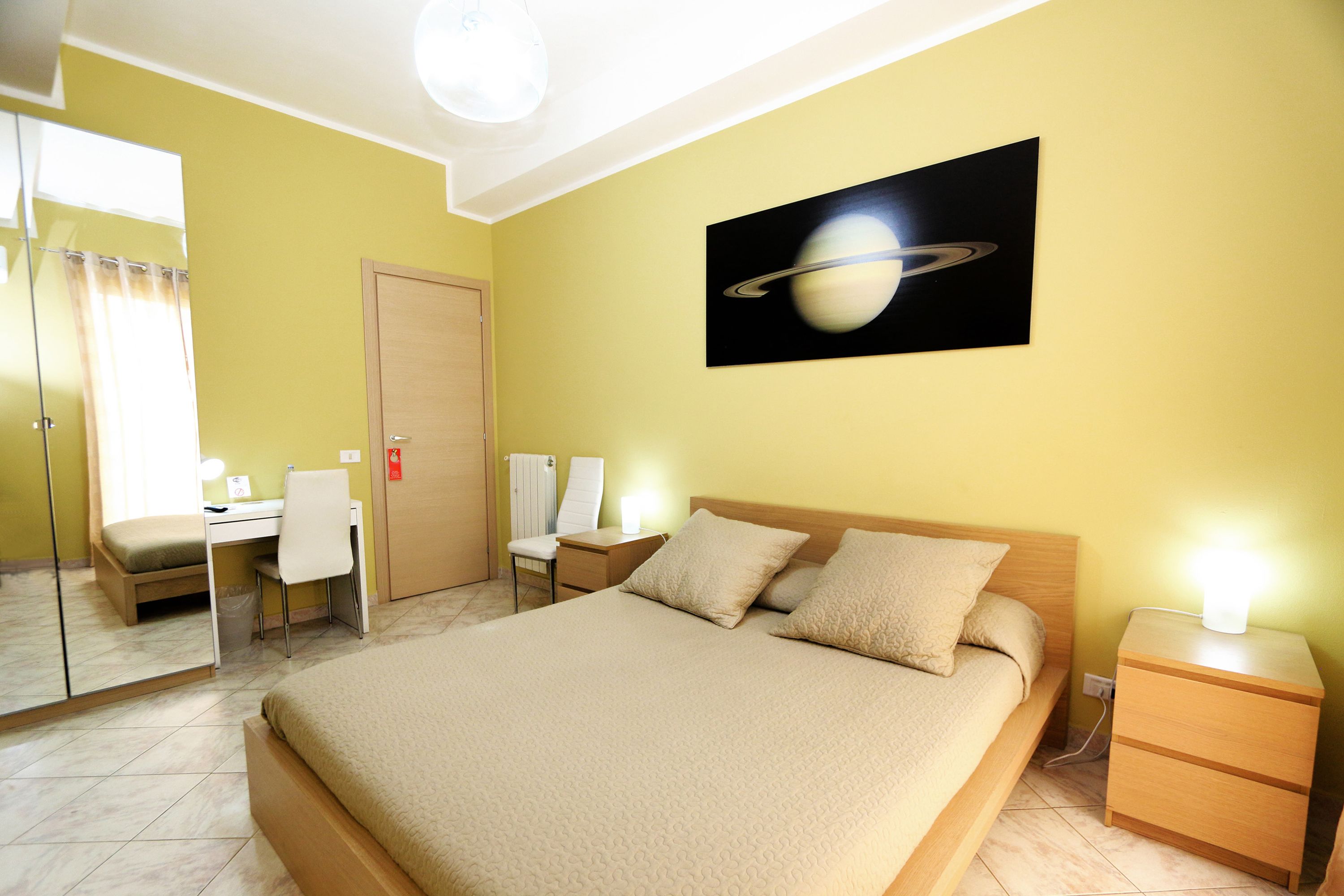 Une chambre d’hôtel comprenant un lit, un bureau, deux chaises, une table de chevet et un tableau représentant Saturne, accroché au mur.
