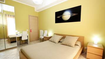 Une chambre d’hôtel comprenant un lit, un bureau, deux chaises, une table de chevet et un tableau représentant Saturne, accroché au mur.
