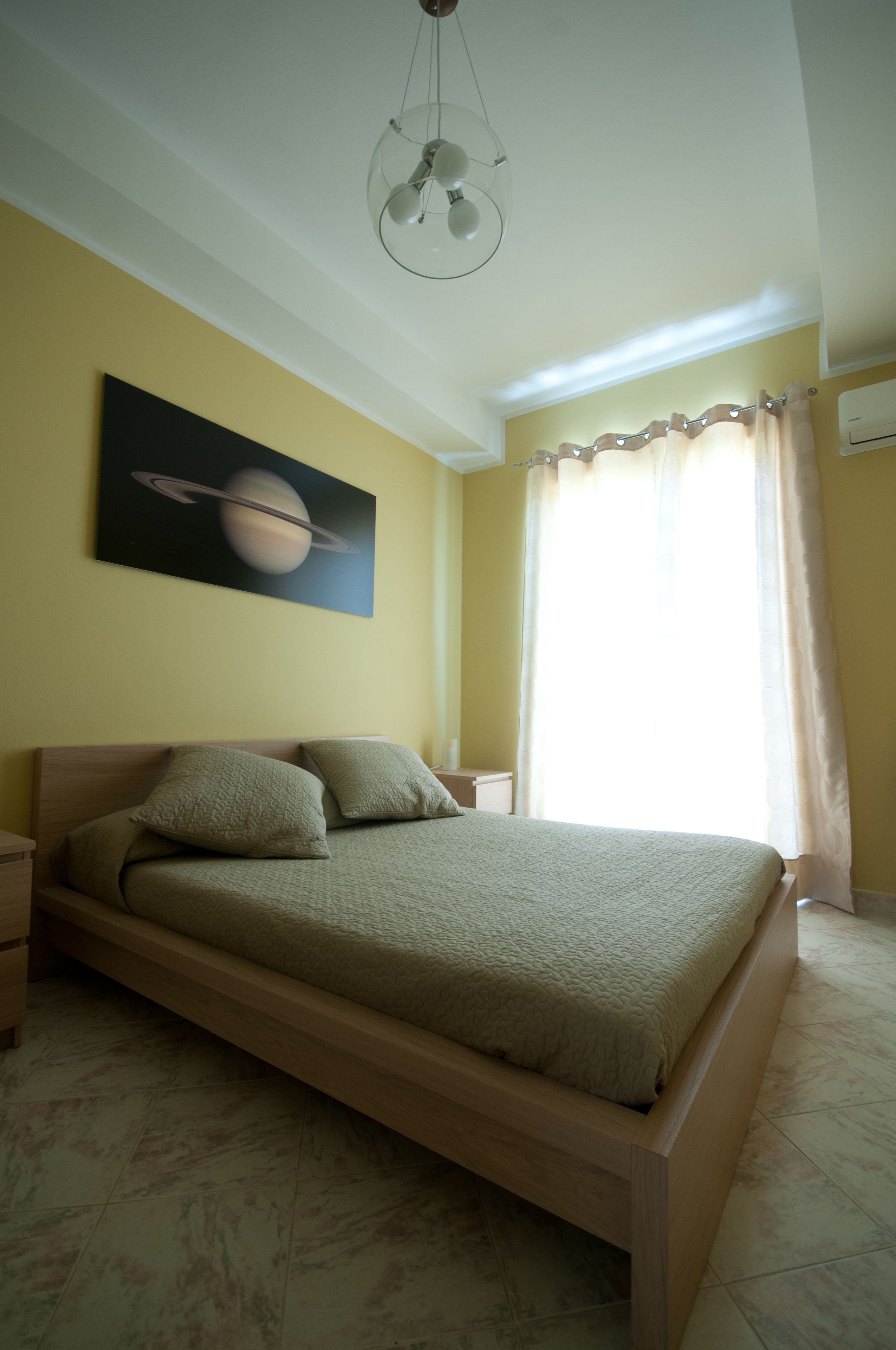Quarto com cama de madeira, mesa de cabeceira, luminária na parede e pintura de Saturno.