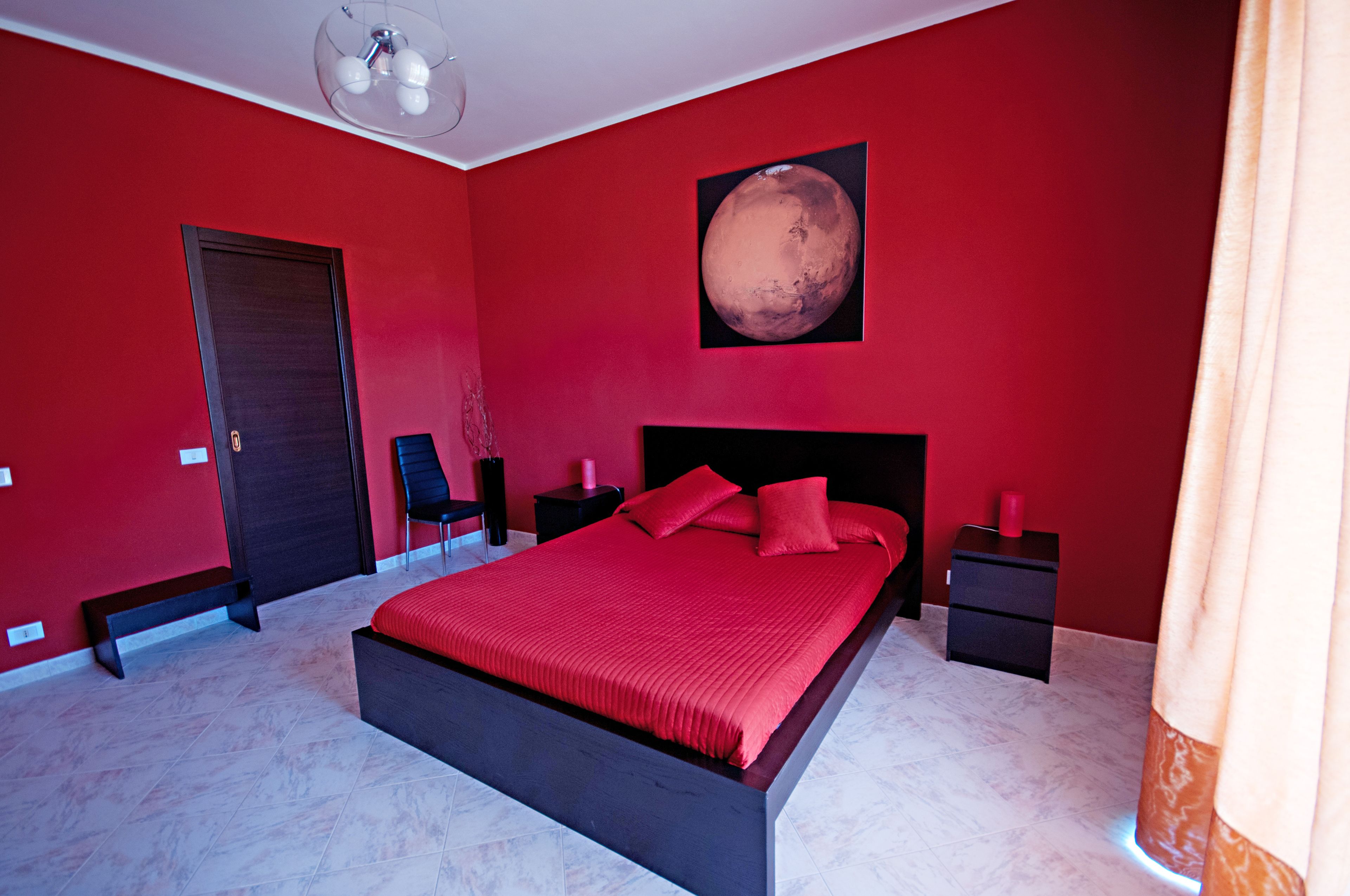 A bedroom with a red wall, a large bed with red bedding, a nightstand, a chair, and a picture of Mars.
