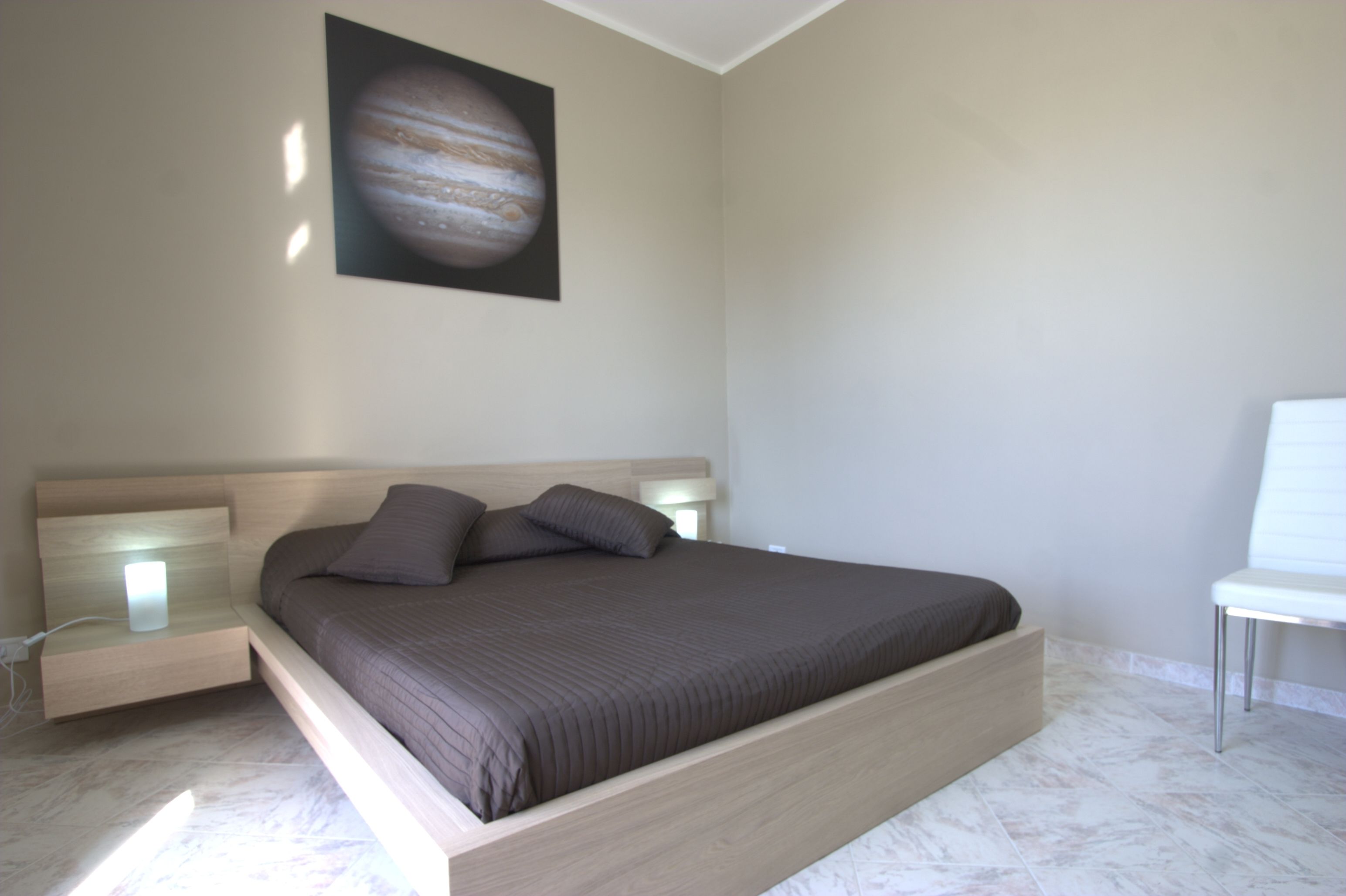 A modern bedroom with a large bed, bedside tables, a wall-mounted artwork of Jupiter, and a white chair.