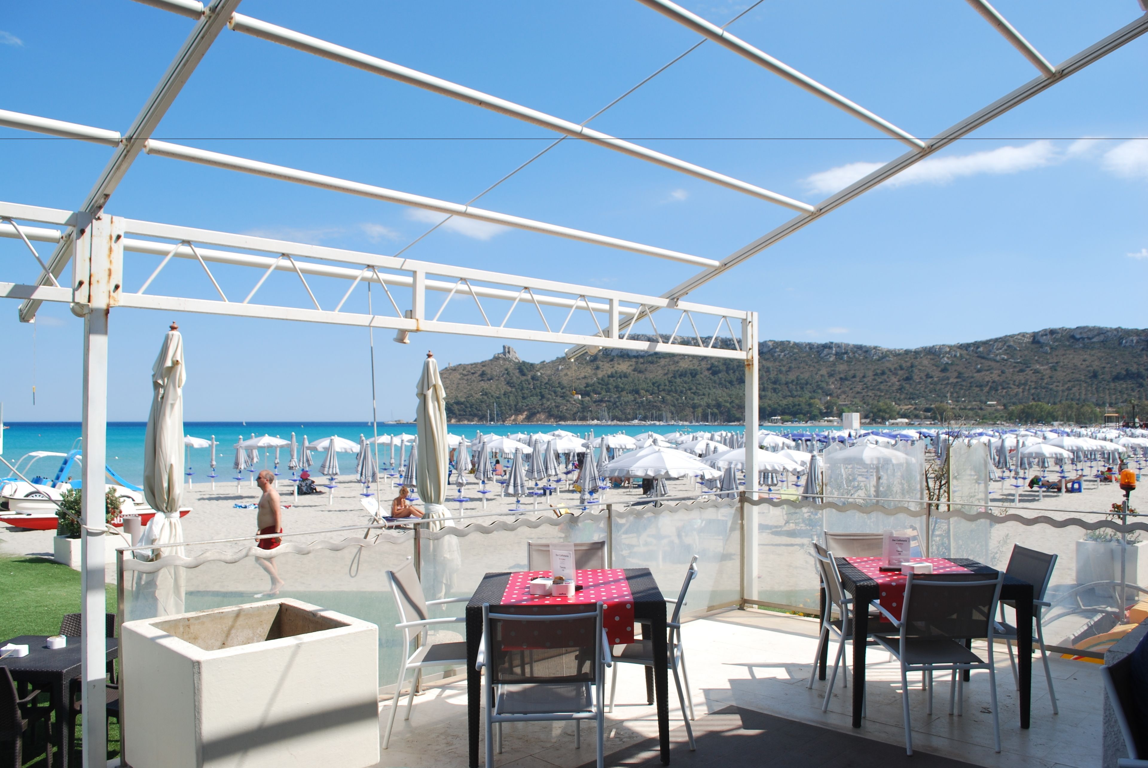 Un espace de restauration en plein air, avec des tables et des chaises, donnant sur une plage parsemée de parasols et fréquentée par des gens.