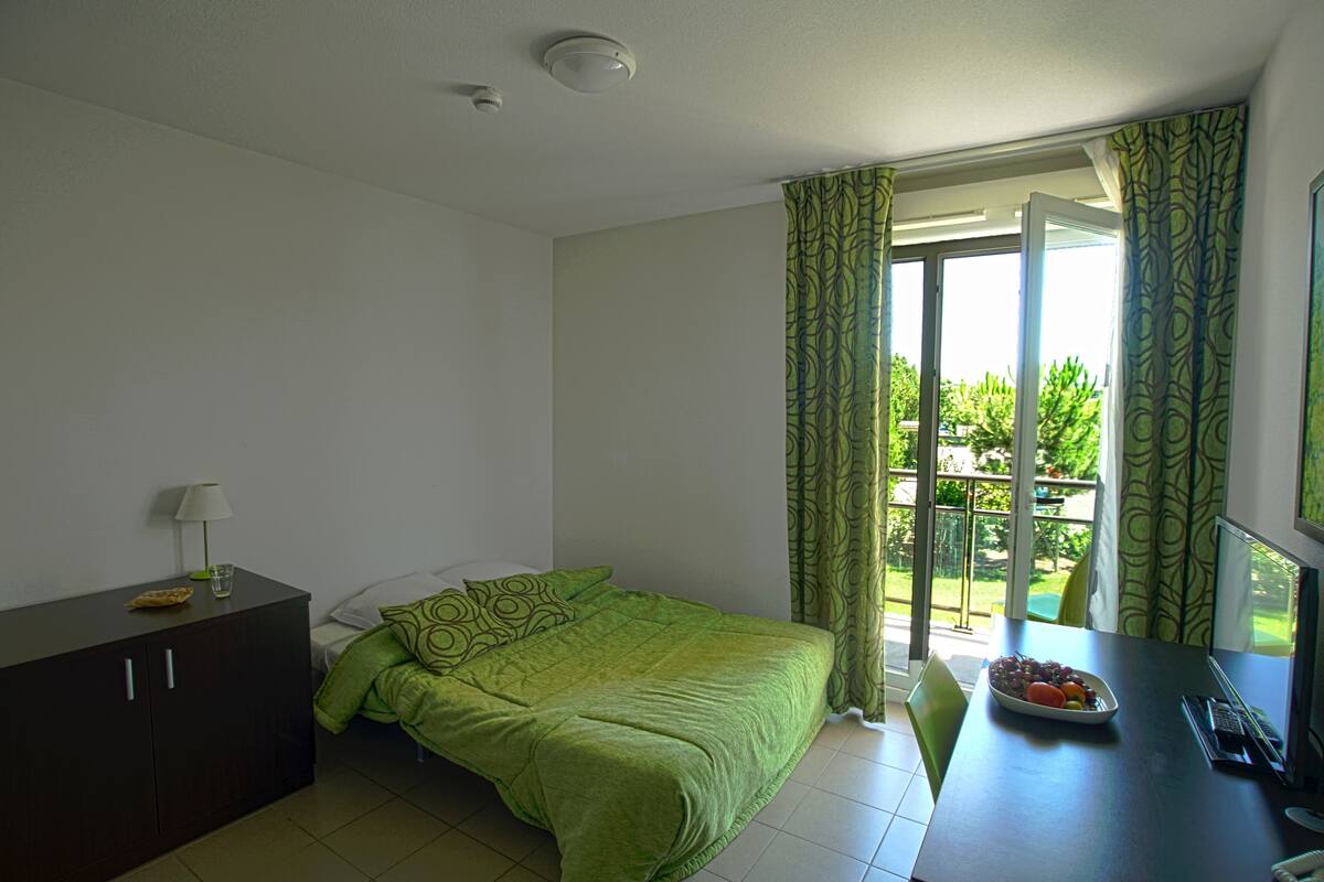 Un dormitorio con una cama, un escritorio con ordenador, una lámpara y una ventana con cortinas.