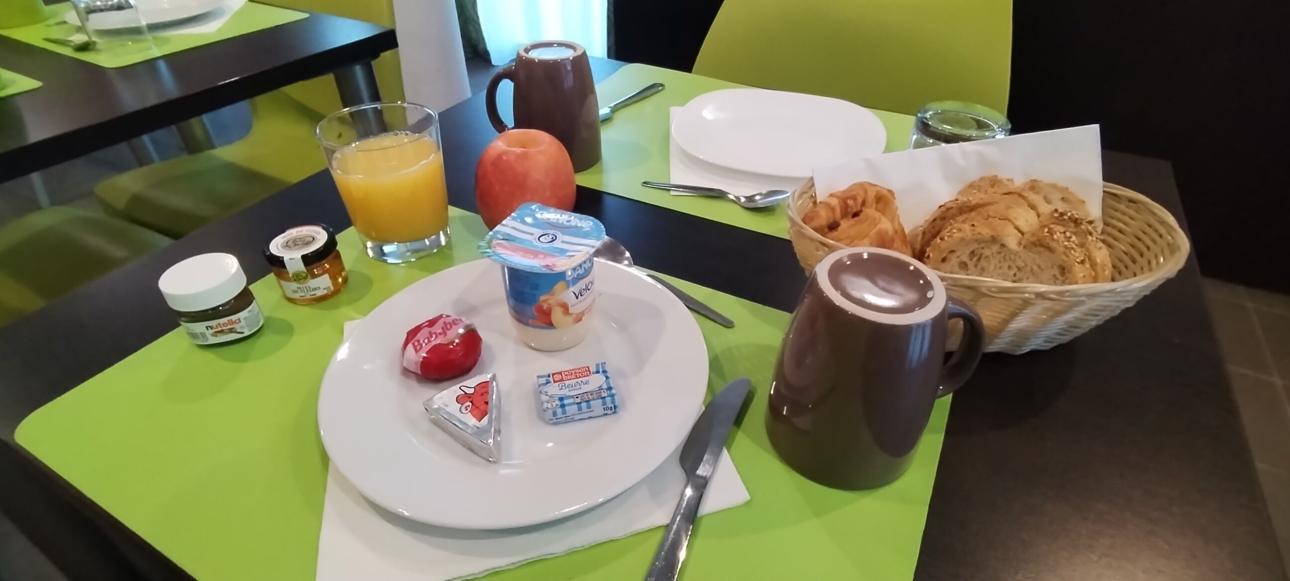 A breakfast table with a plate, juice, a mug, a basket of croissants, and a bottle of milk.
