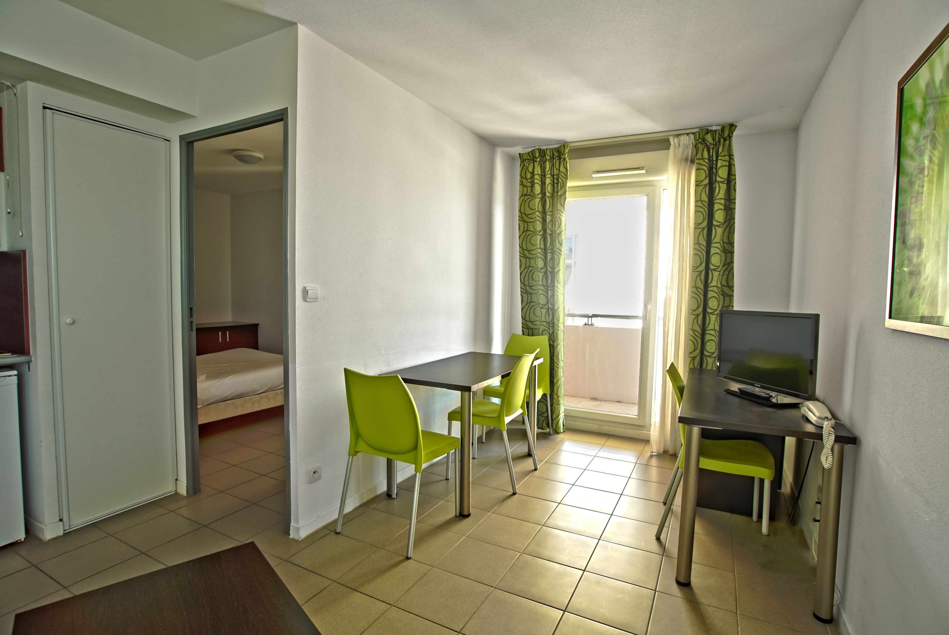 Una habitación pequeña con cama, una mesa de comedor con sillas, televisor y ventana con cortinas.