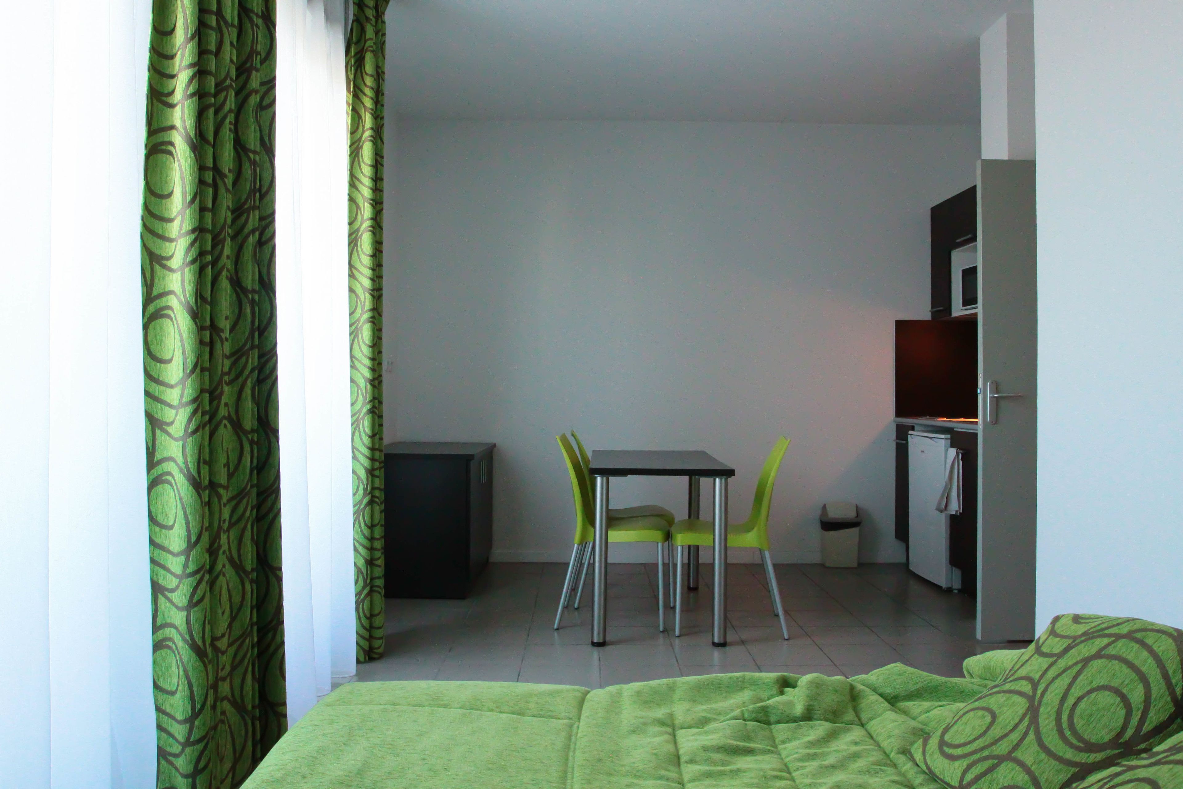 A small, modern room with a bed, a desk, two chairs, a small kitchenette, and a trash bin.