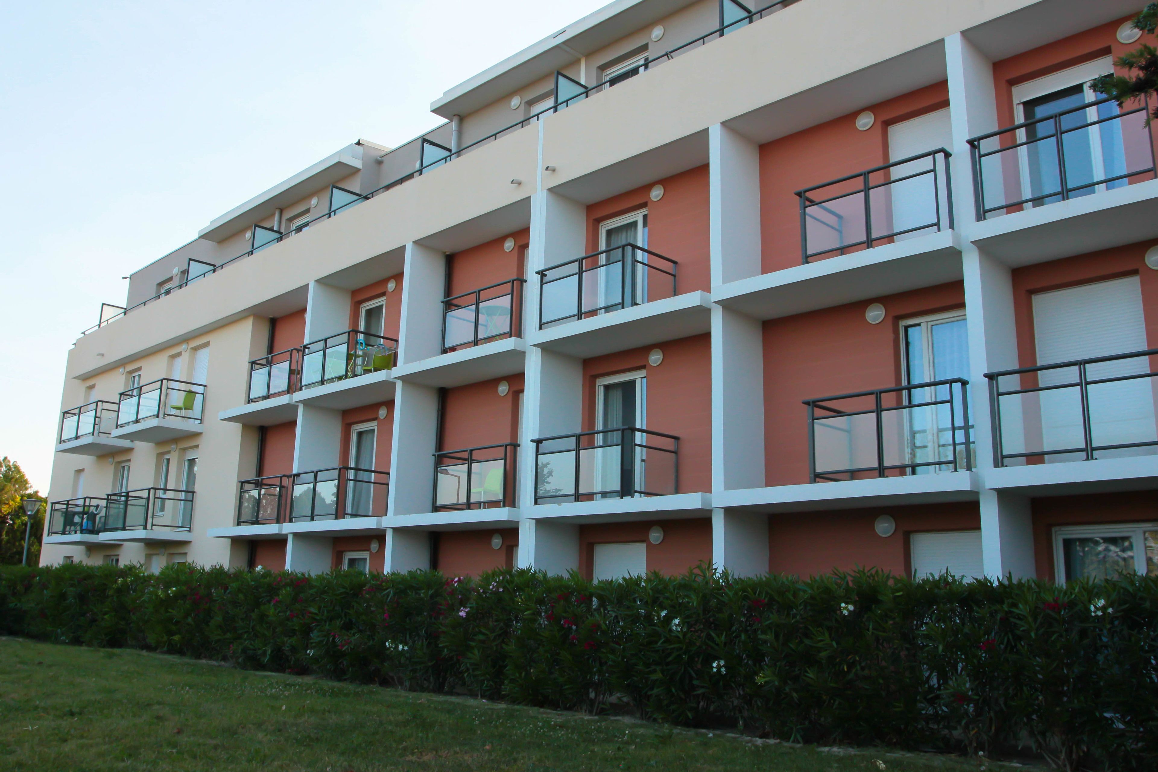 A multi-story residential building with balconies, a flat roof, and a well-maintained hedge in front.