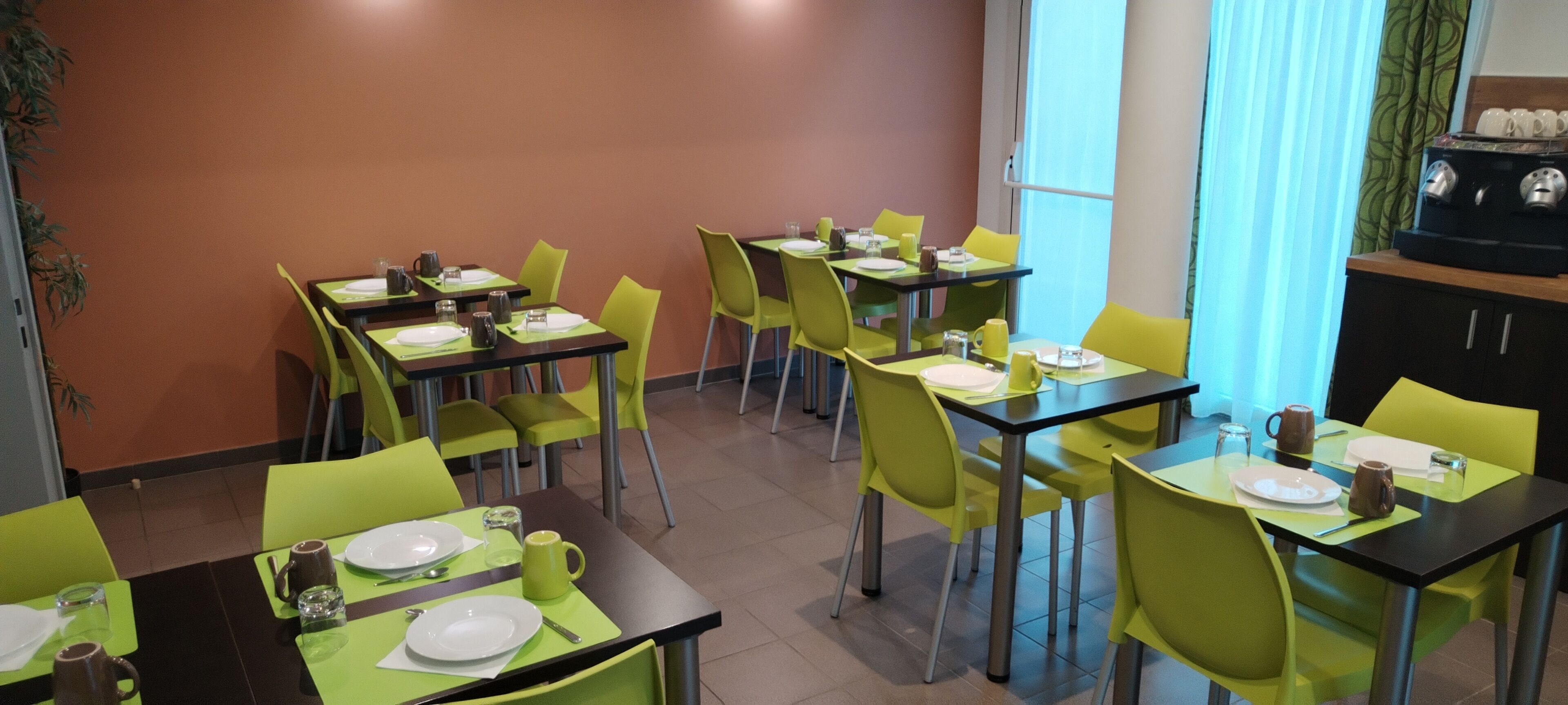 Un comedor con mesas y sillas, una cafetera y un ventanal con cortinas.