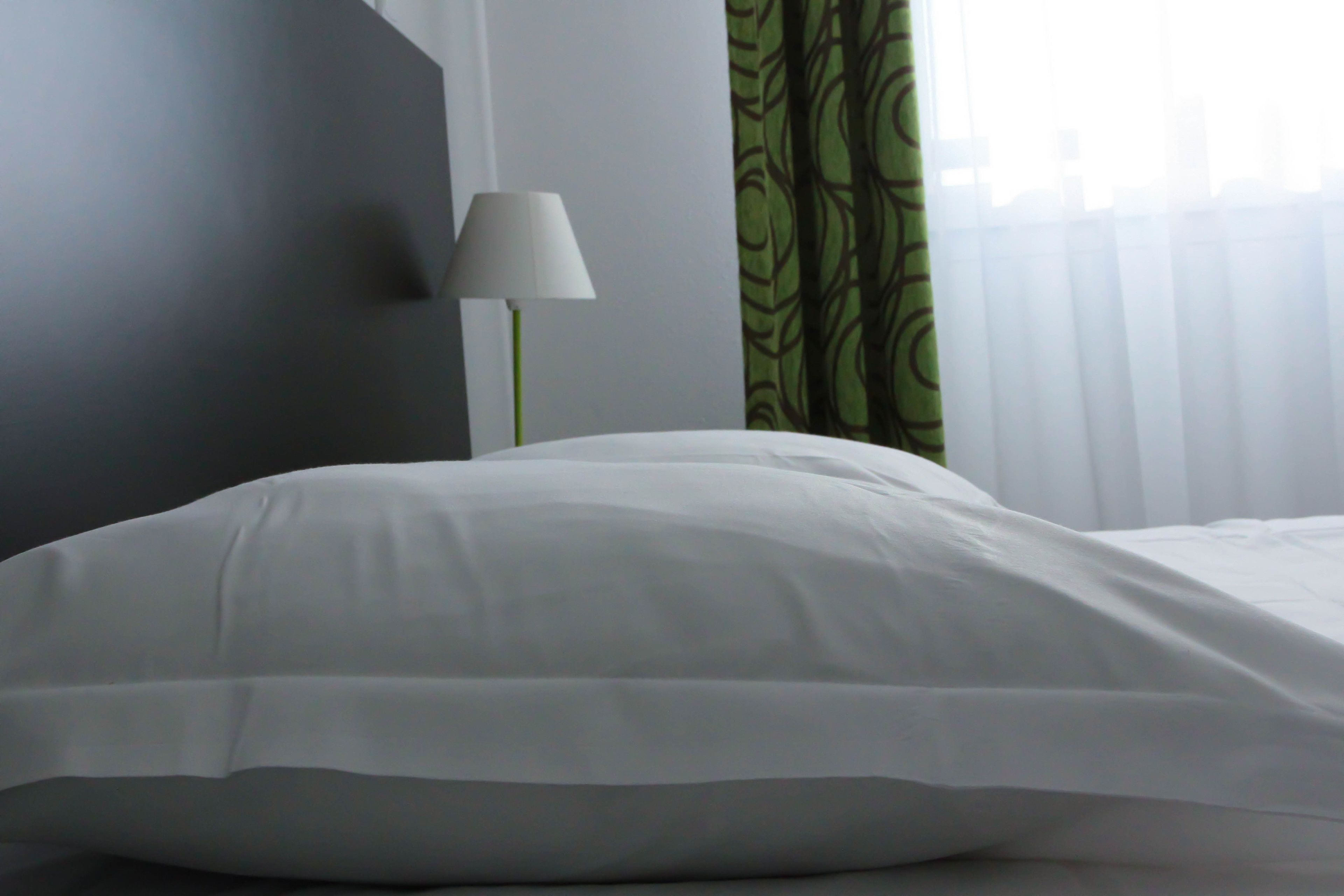 Un llit ben fet amb roba de llit blanca en una habitació amb una cortina verda amb dibuixos i una làmpada de peu.