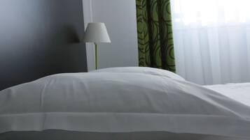 Un llit ben fet amb roba de llit blanca en una habitació amb una cortina verda amb dibuixos i una làmpada de peu.