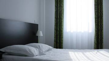 A neatly made bed with white linens in a room with a dark headboard, a bedside lamp, and sheer green curtains over a window.