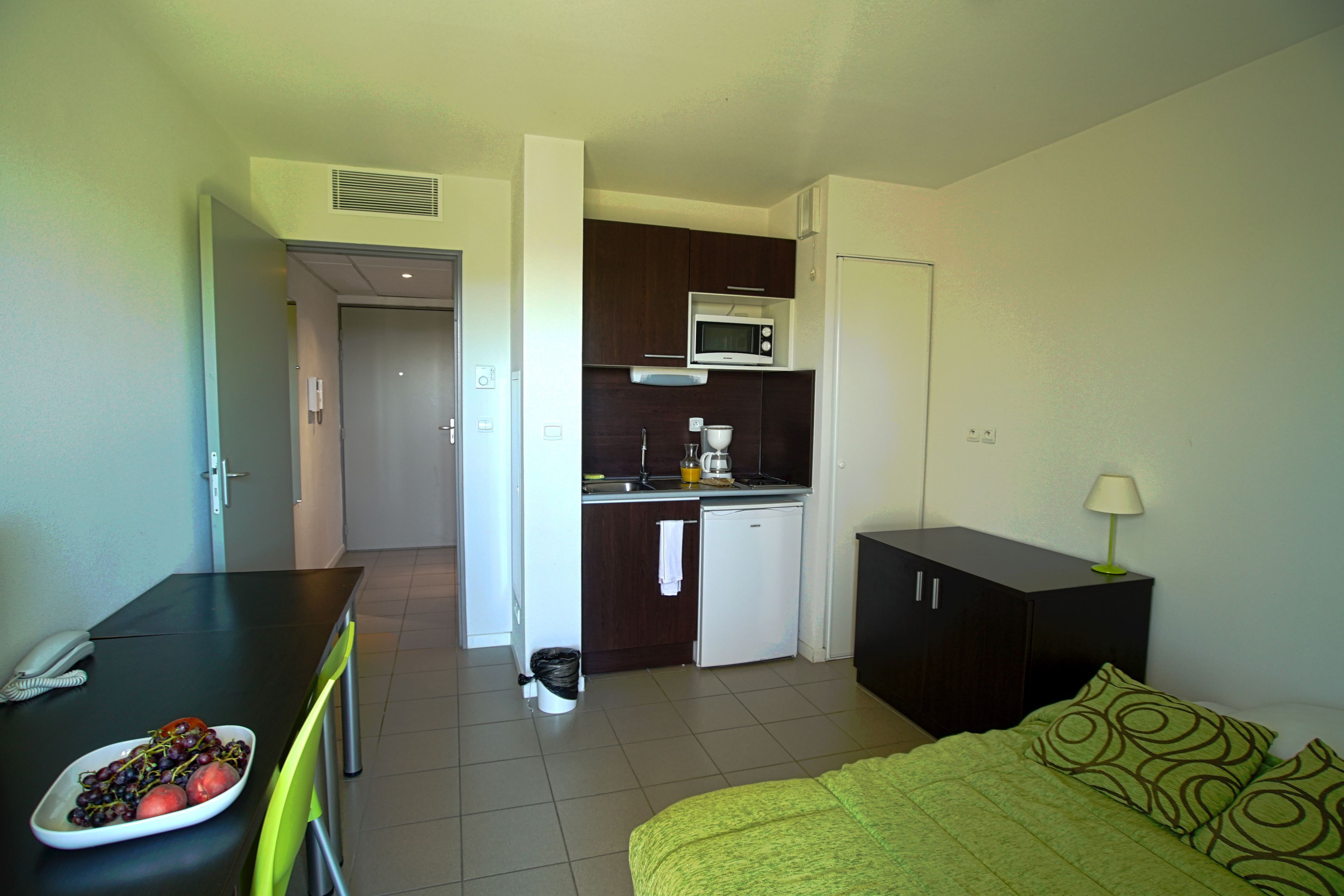 Habitación de hotel compacta con kitchenette, una cama con ropa de cama verde, un escritorio con un plato de fruta y una mesita de noche pequeña con una lámpara.