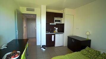 Habitación de hotel compacta con kitchenette, una cama con ropa de cama verde, un escritorio con un plato de fruta y una mesita de noche pequeña con una lámpara.