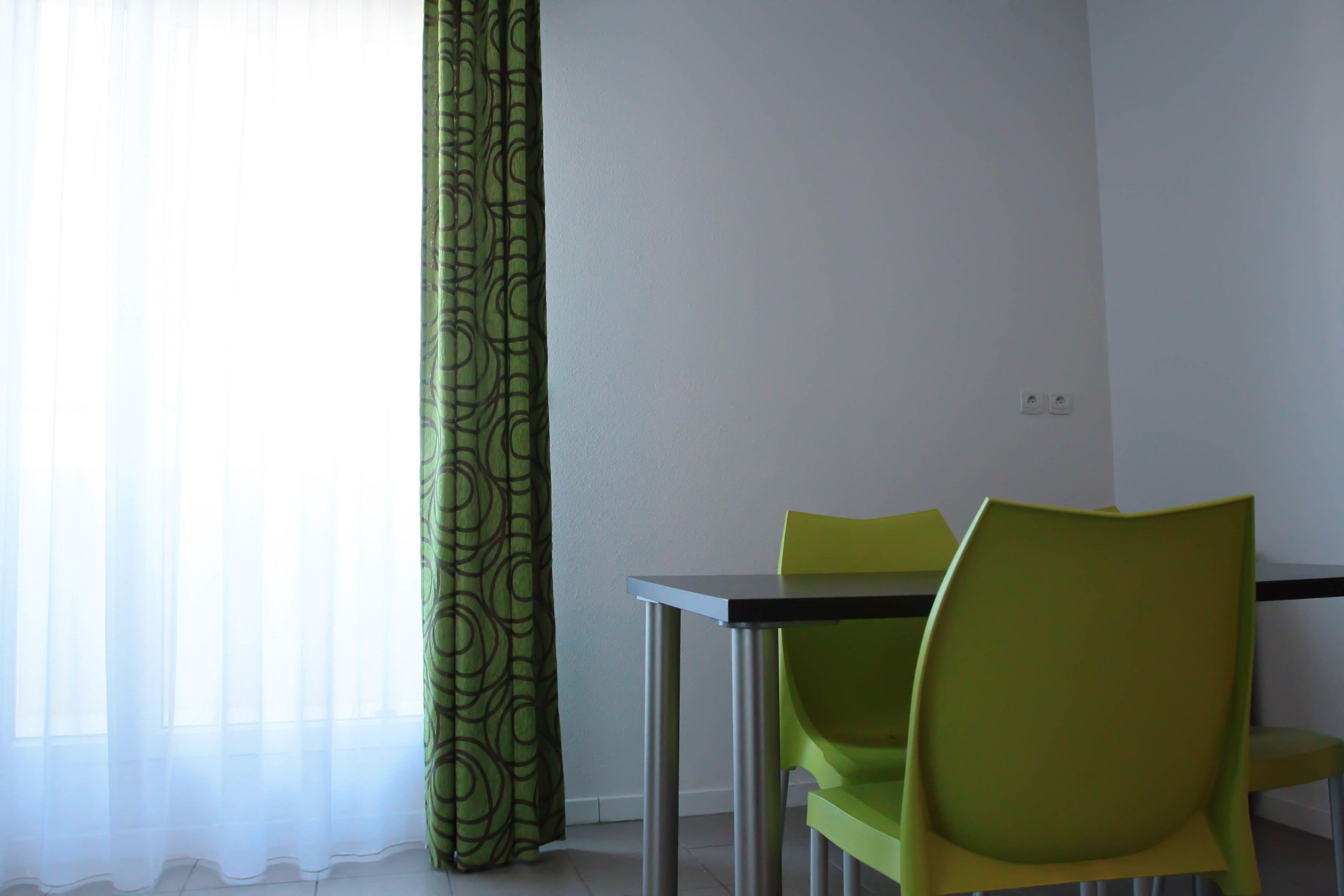 Un coin repas avec une table et deux chaises, une fenêtre avec des rideaux légers et un mur sobre agrémenté d’un interrupteur.