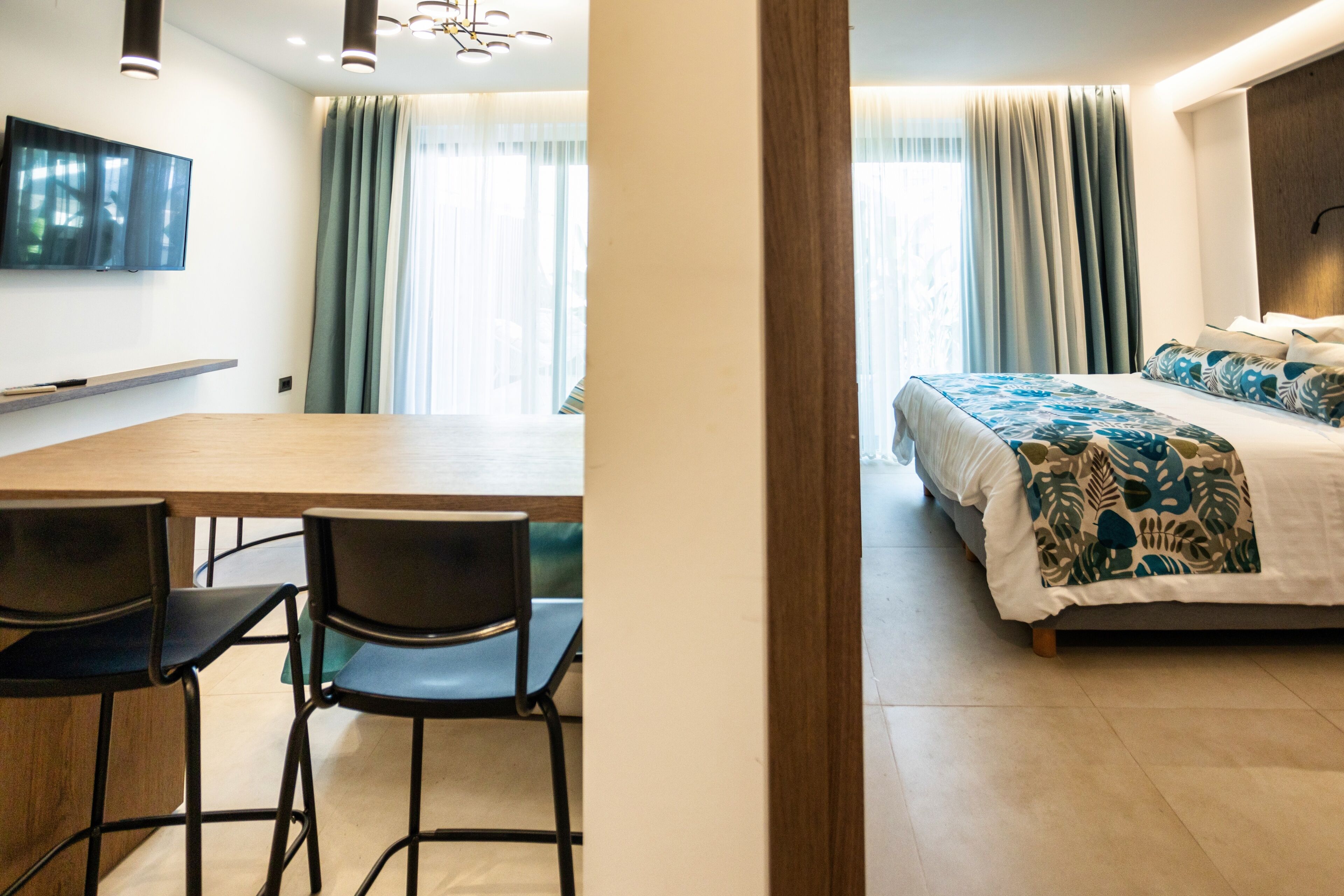 Une chambre d’hôtel moderne équipée d’un lit, d’un téléviseur fixé au mur, d’une table à manger avec des chaises et d’une grande fenêtre avec des rideaux.