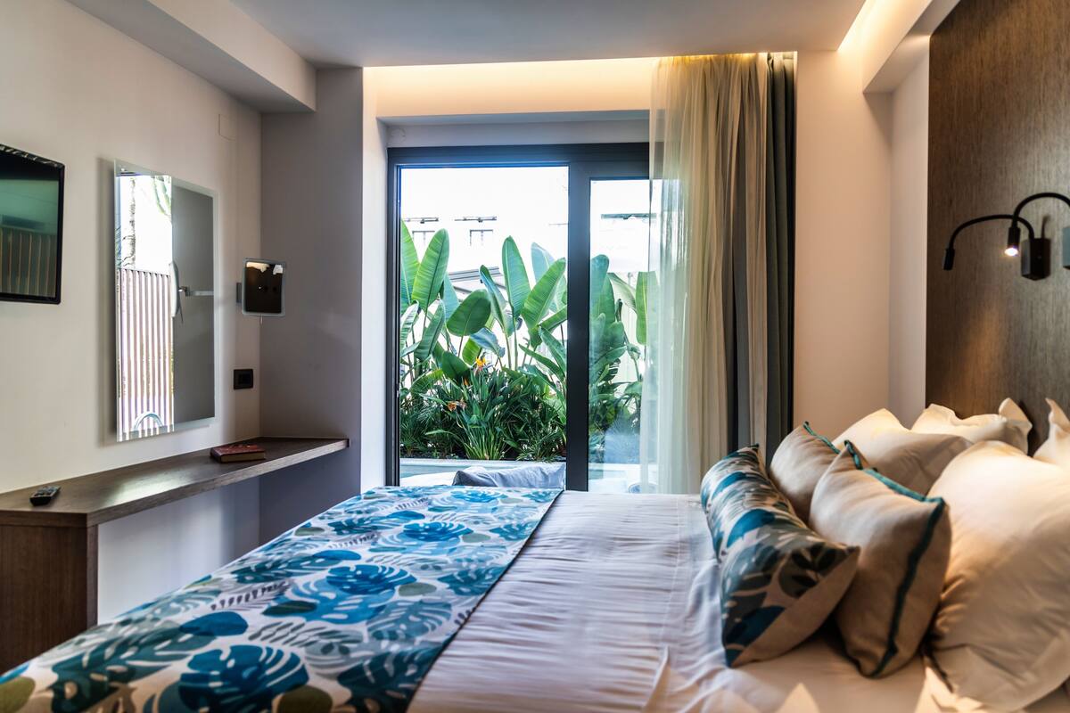 A bedroom with a bed, a bedside table, a mirror, a window with curtains, and a view of greenery outside.