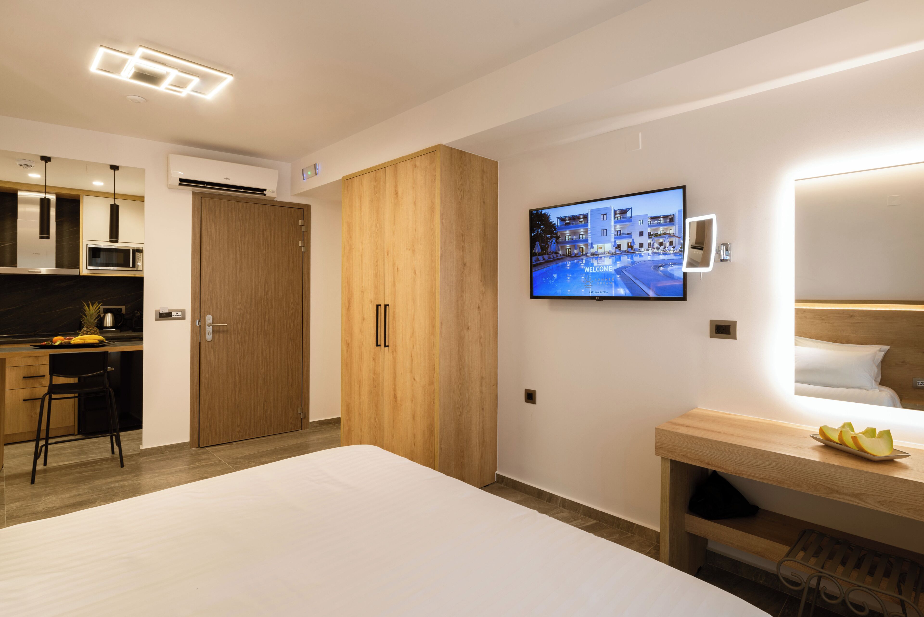 A modern hotel room with a bed, a TV mounted on the wall, a wooden wardrobe, and a kitchenette area.