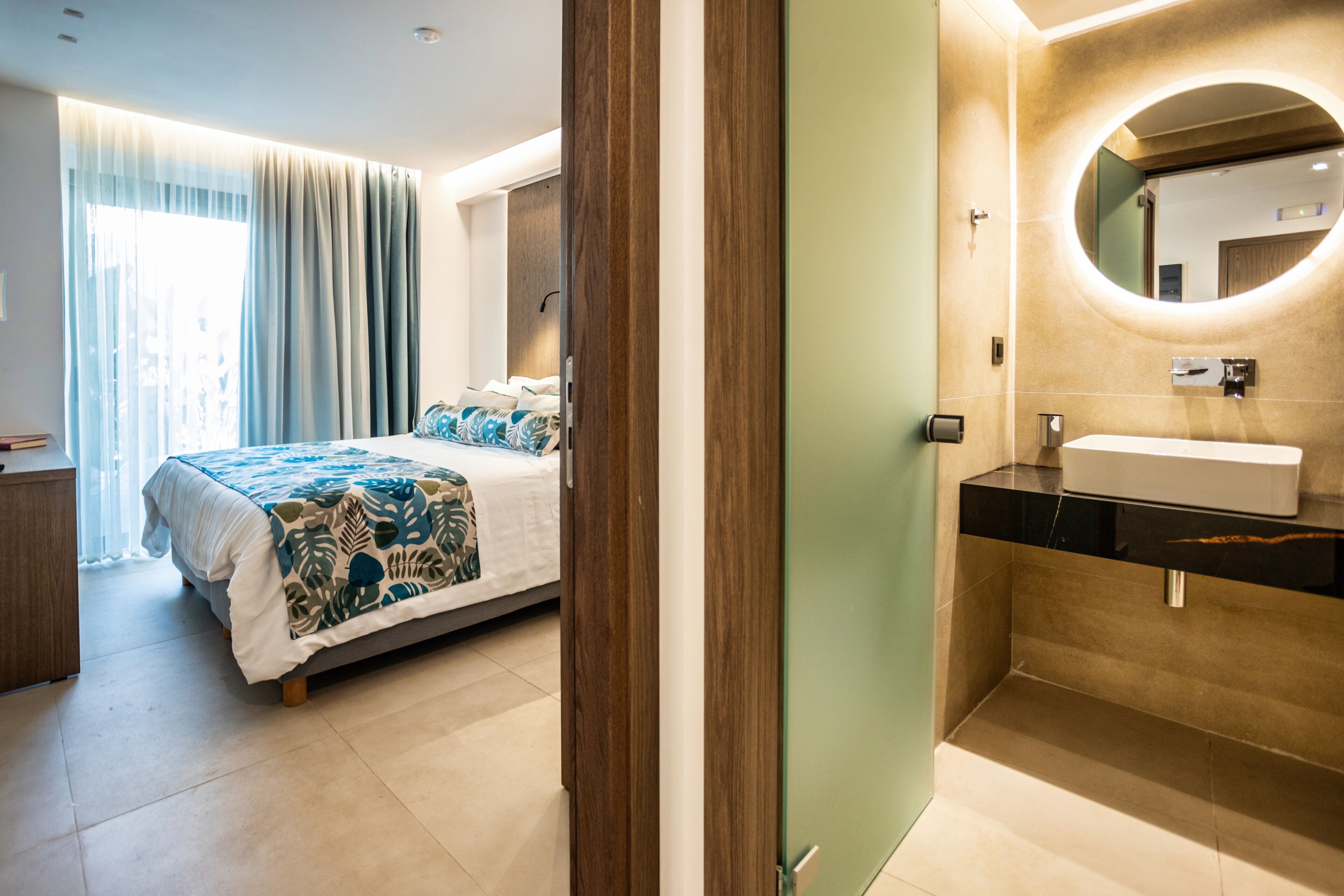 A modern hotel room with a bed, a bathroom with a sink and mirror, and a view of the outside.