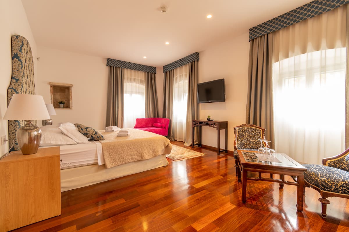 Ein Hotelzimmer mit Holzboden, einem Bett mit weißen Leinentüchern, einem roten Sessel, einem Holztisch, einem an der Wand montierten Fernseher und einem Fenster mit Vorhängen.