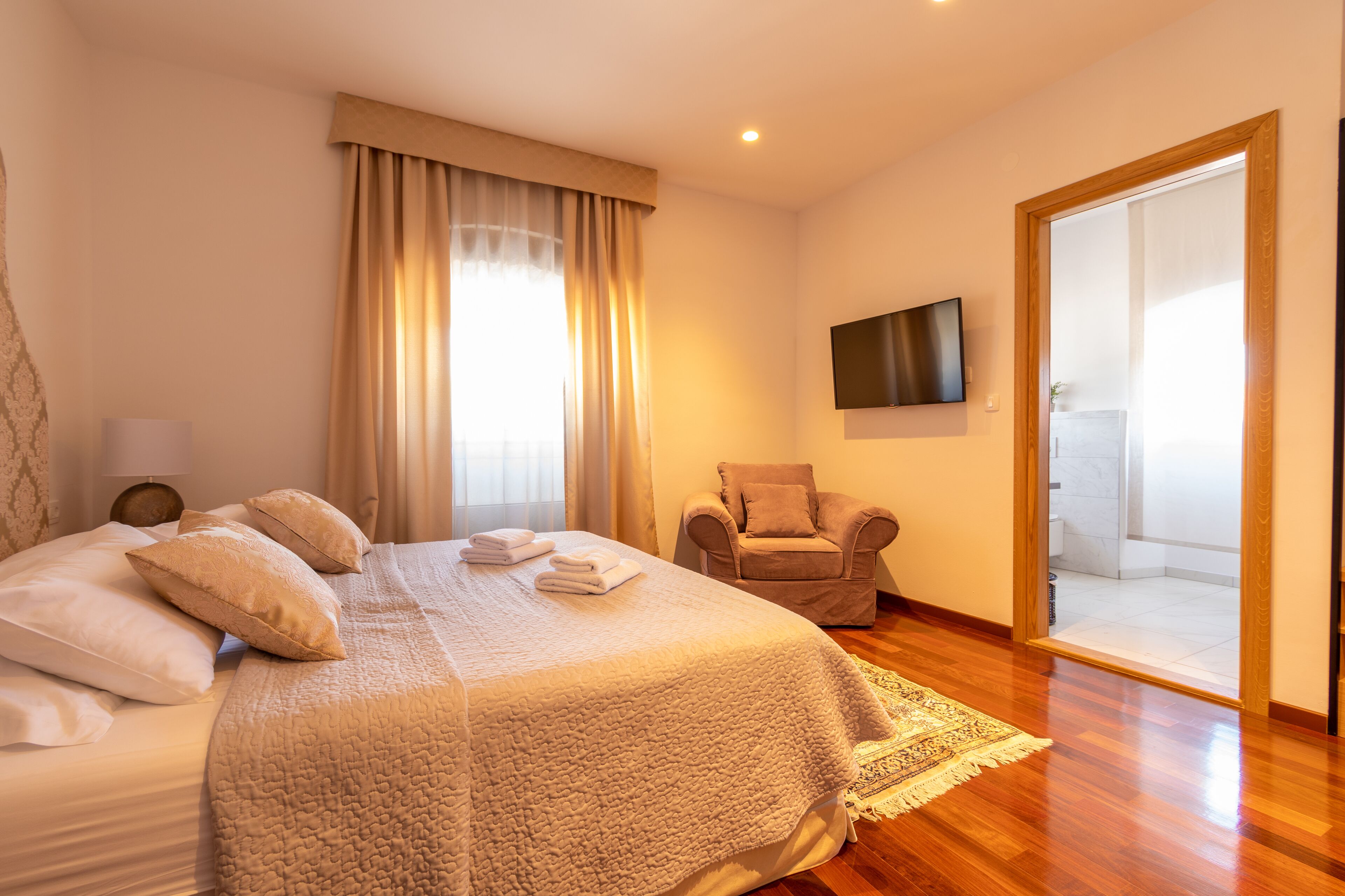 A hotel room with a large bed, a chair, a TV, and a bathroom visible through an open door.