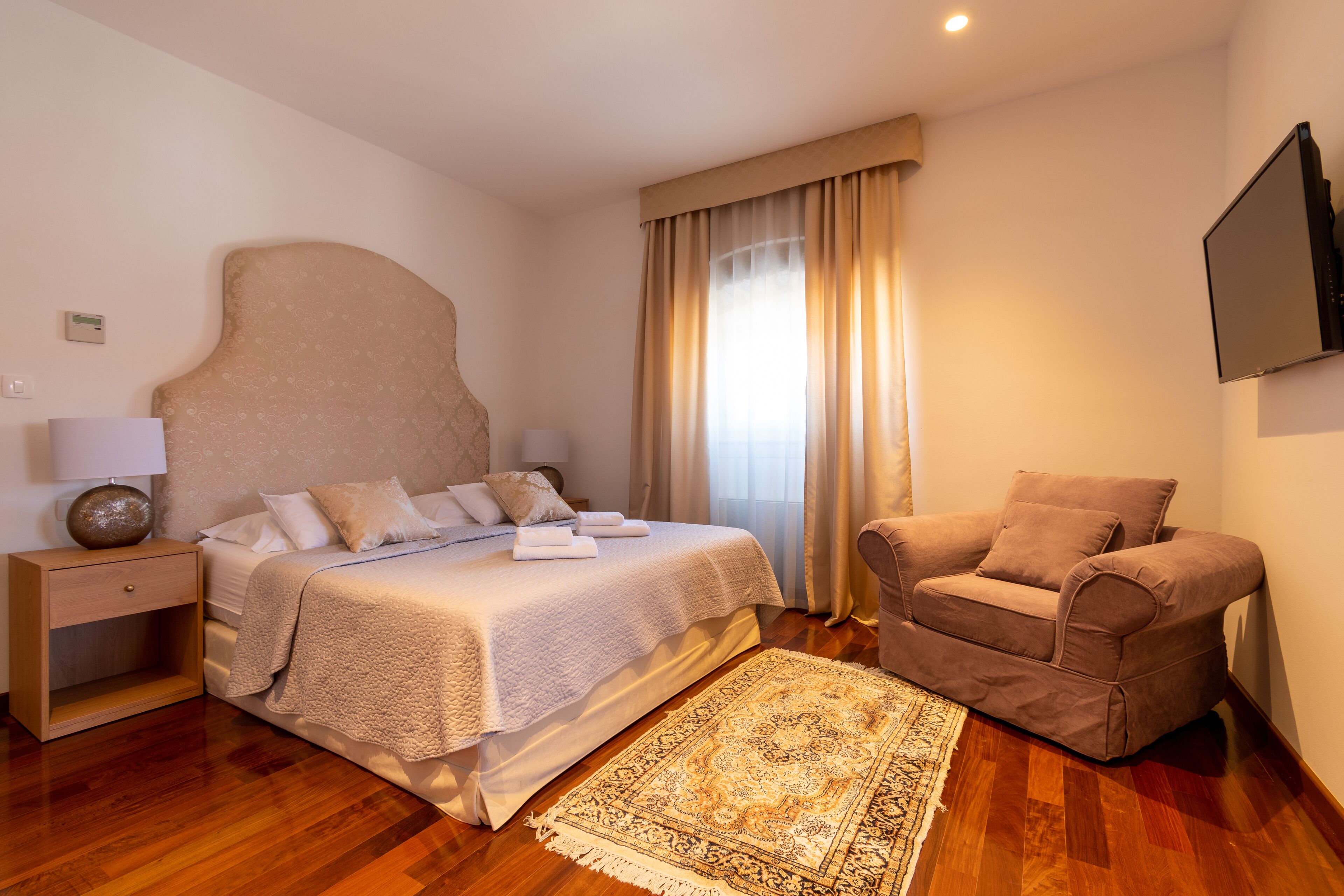 A bedroom with a large bed, a nightstand, a sofa, and a TV mounted on the wall.