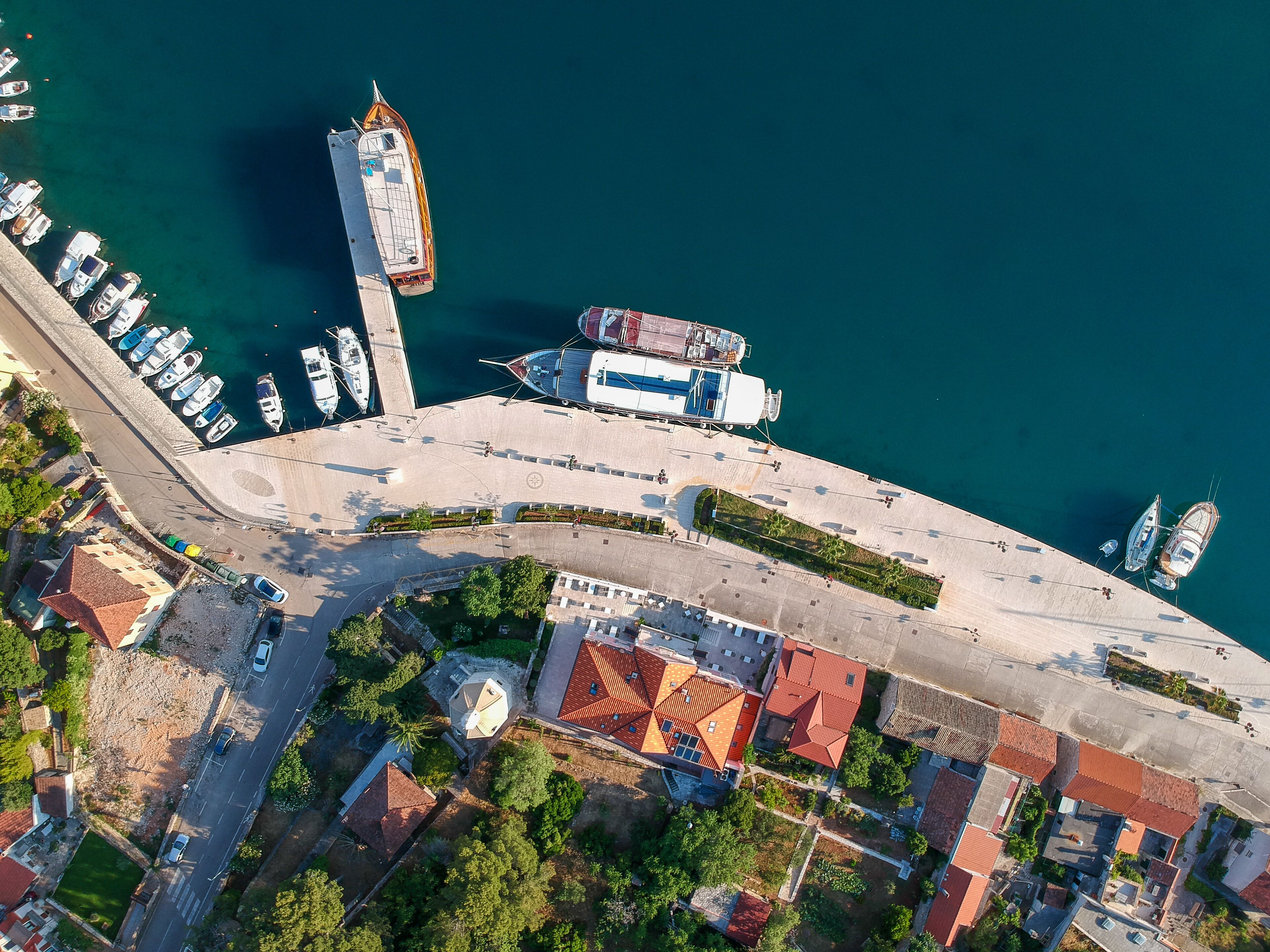 An aerial view of a coastal marina with boats docked, a large white building, and residential buildings with red roofs.