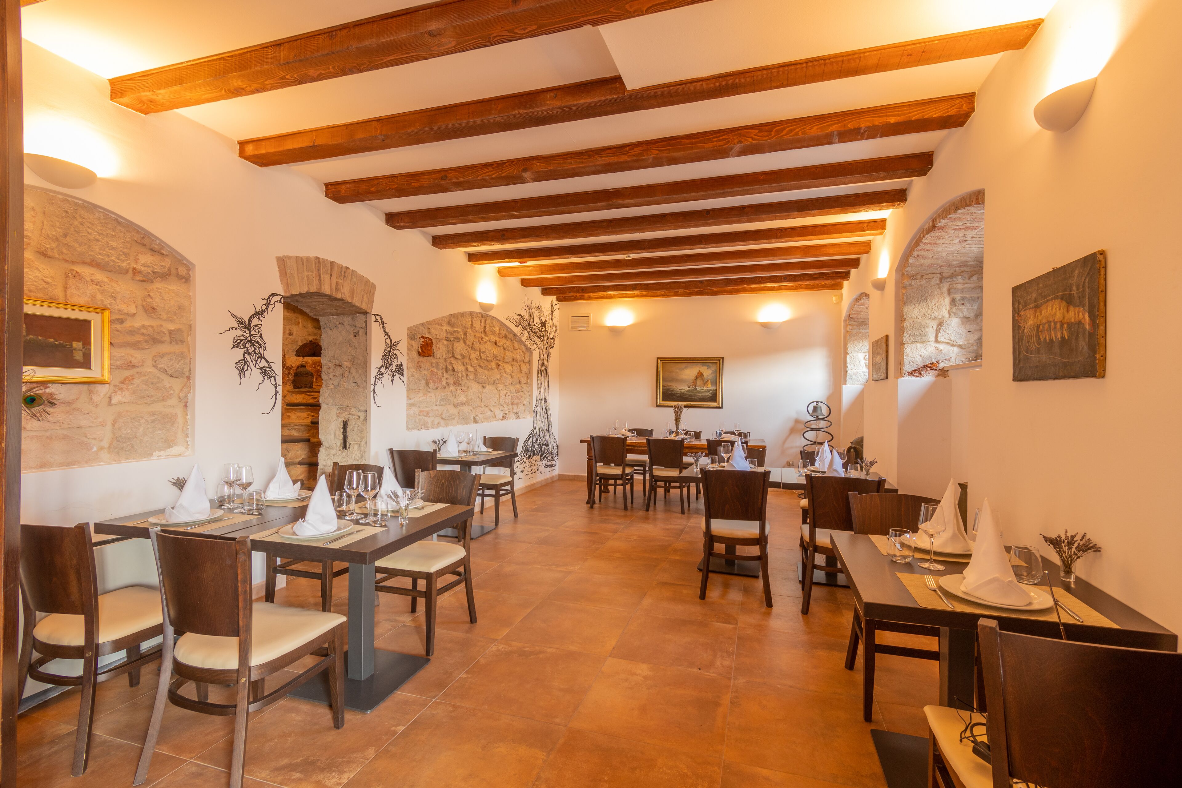 Un restaurant doté de poutres en bois, de murs en pierre et de nombreuses tables, toutes recouvertes de nappes blanches et entourées de chaises.