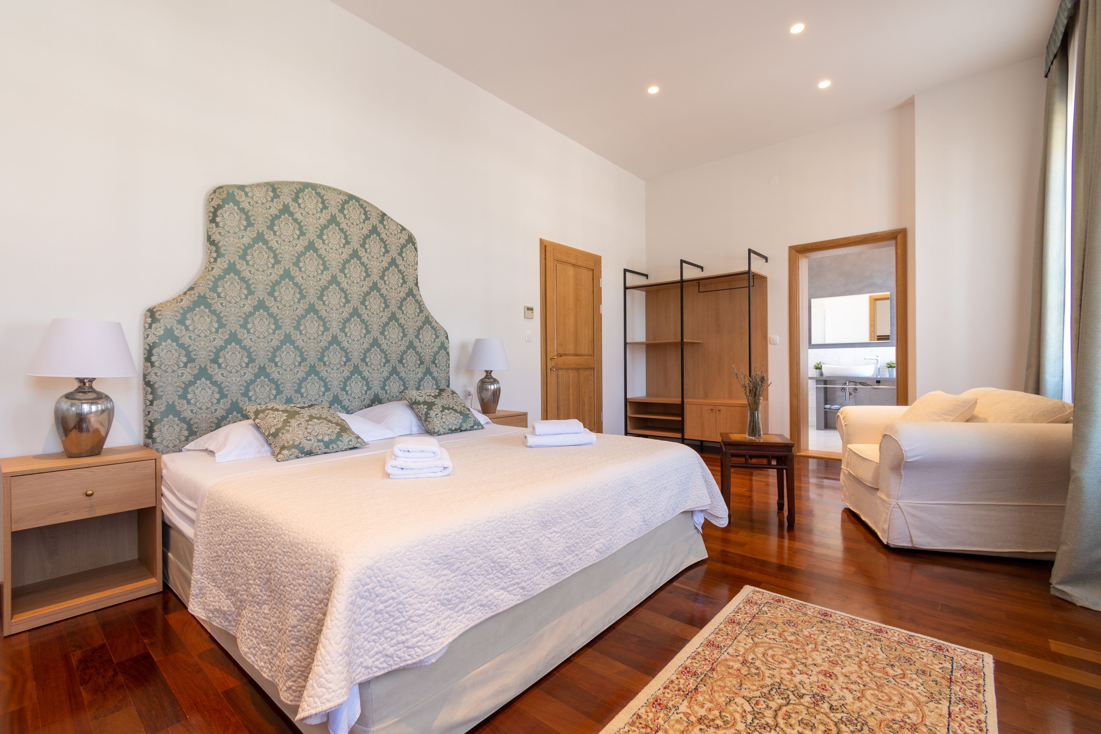 Una habitación de hotel con una cama grande, una mesita de noche de madera, una mesita de noche con lámparas, un armario, un sofá y un espejo.
