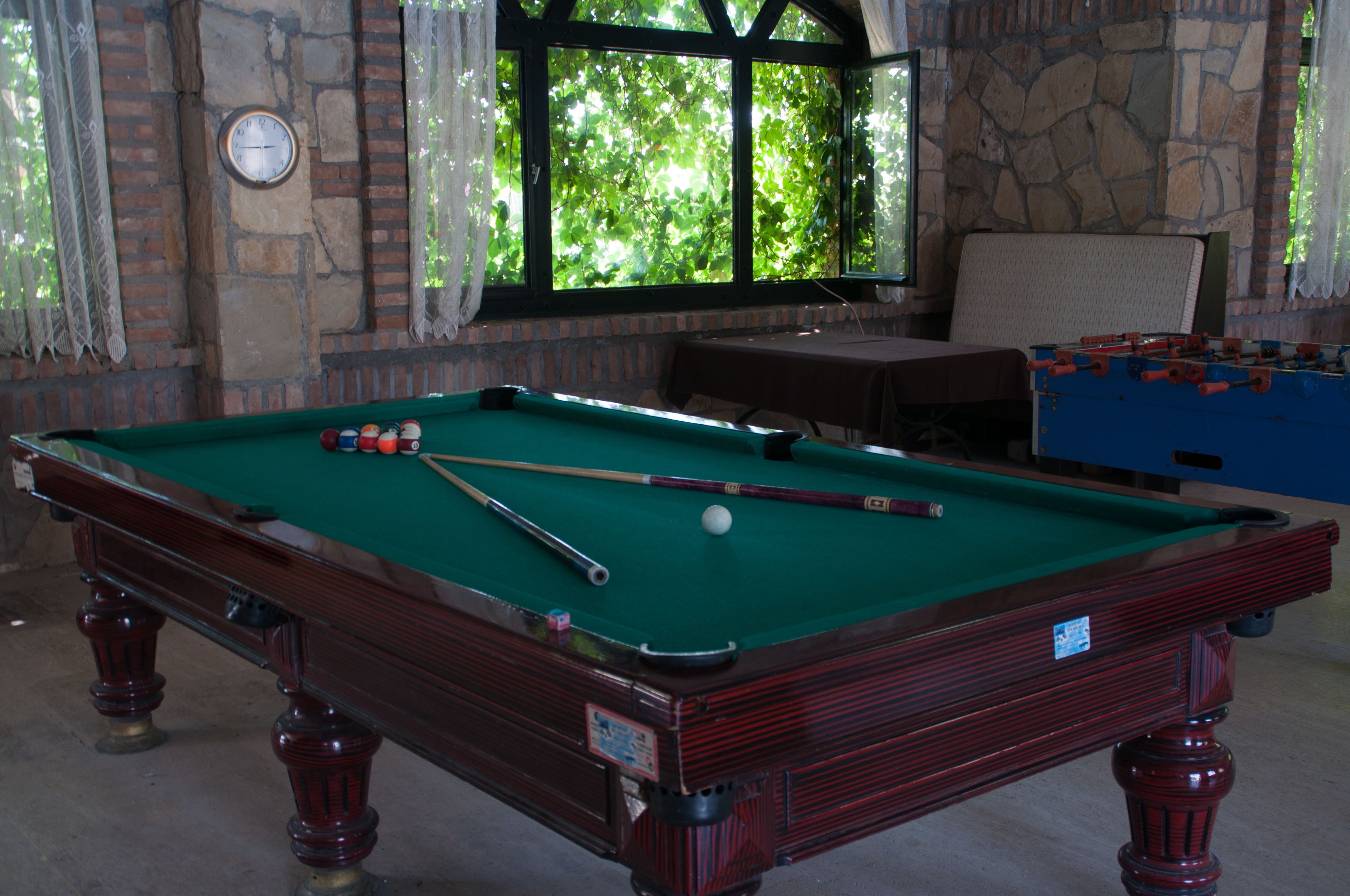 A pool table with balls and cues, a clock on the wall, and a window with a view of greenery.