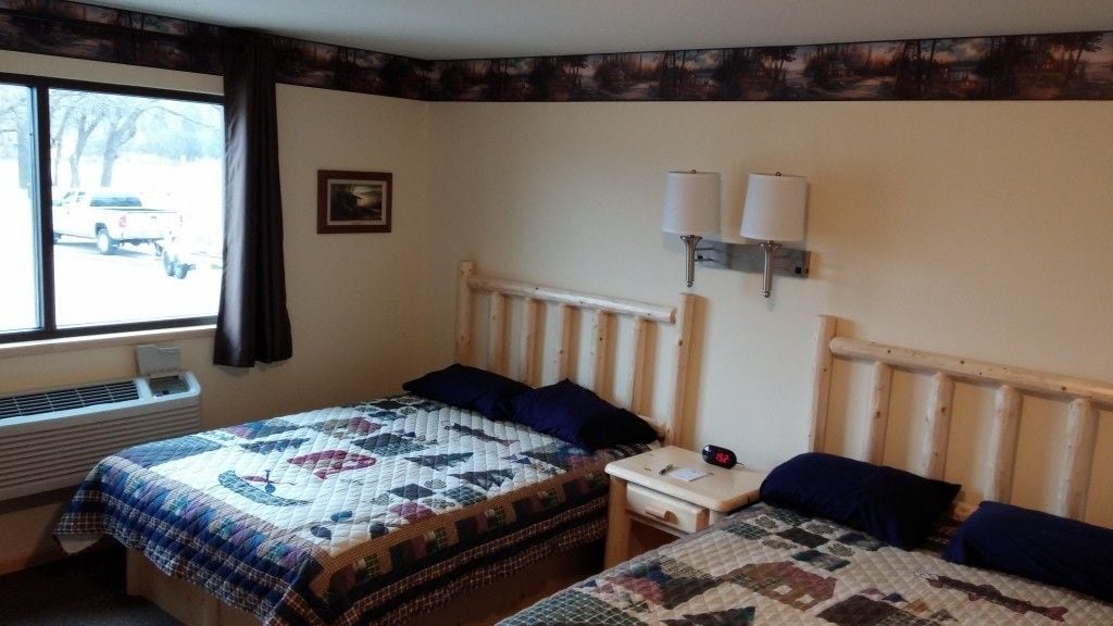 Ein Hotelzimmer mit einem großen Bett, einem Einzelbett, zwei Nachttischen, einem Fenster mit Vorhängen, einer Wandleuchte und einem Bild an der Wand.
