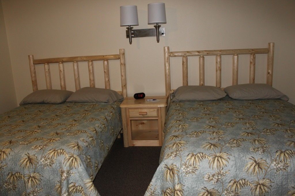 A room with two single beds, each with a wooden headboard and a floral bedspread. There are two wall-mounted lamps above the beds and a small wooden nightstand with a clock between them.